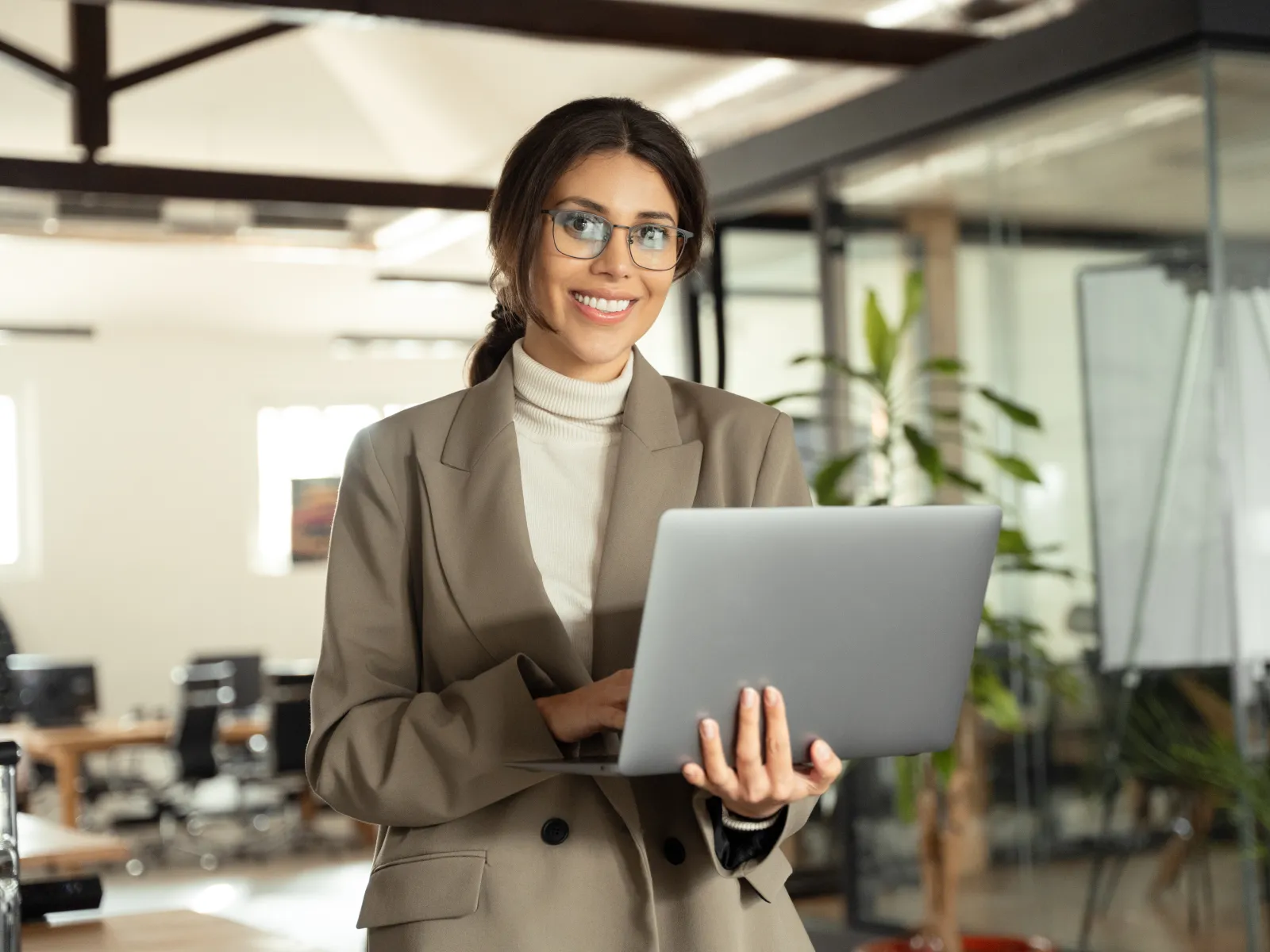 Smiling businesswoman in glasses using laptop in modern office with glass walls and plants.