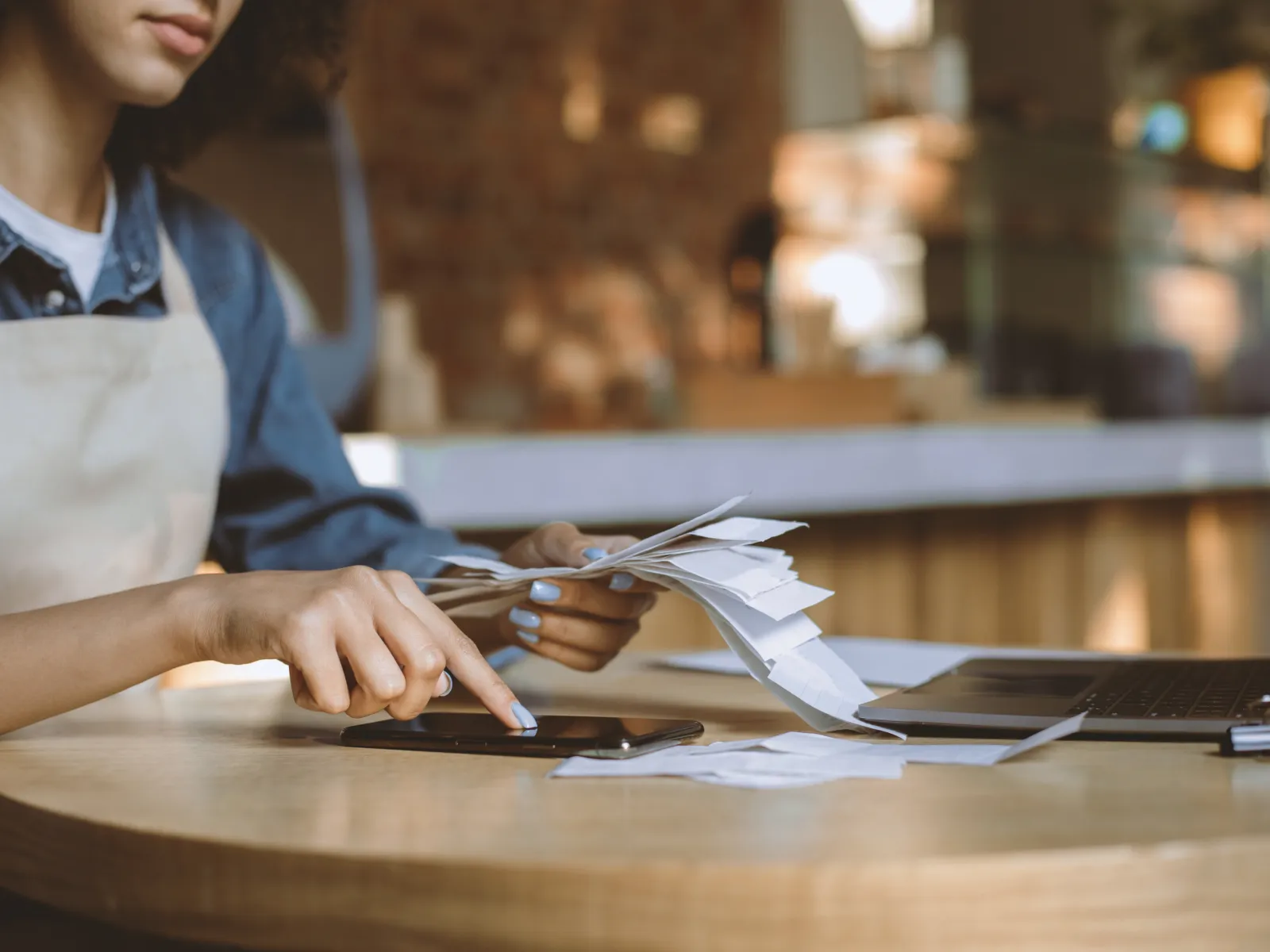 Woman in apron managing receipts and using smartphone at round wooden table with laptop.