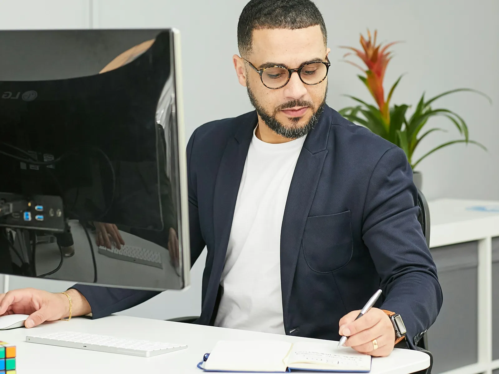 Man in glasses and blazer writing notes at a desk with computer and plants in modern office