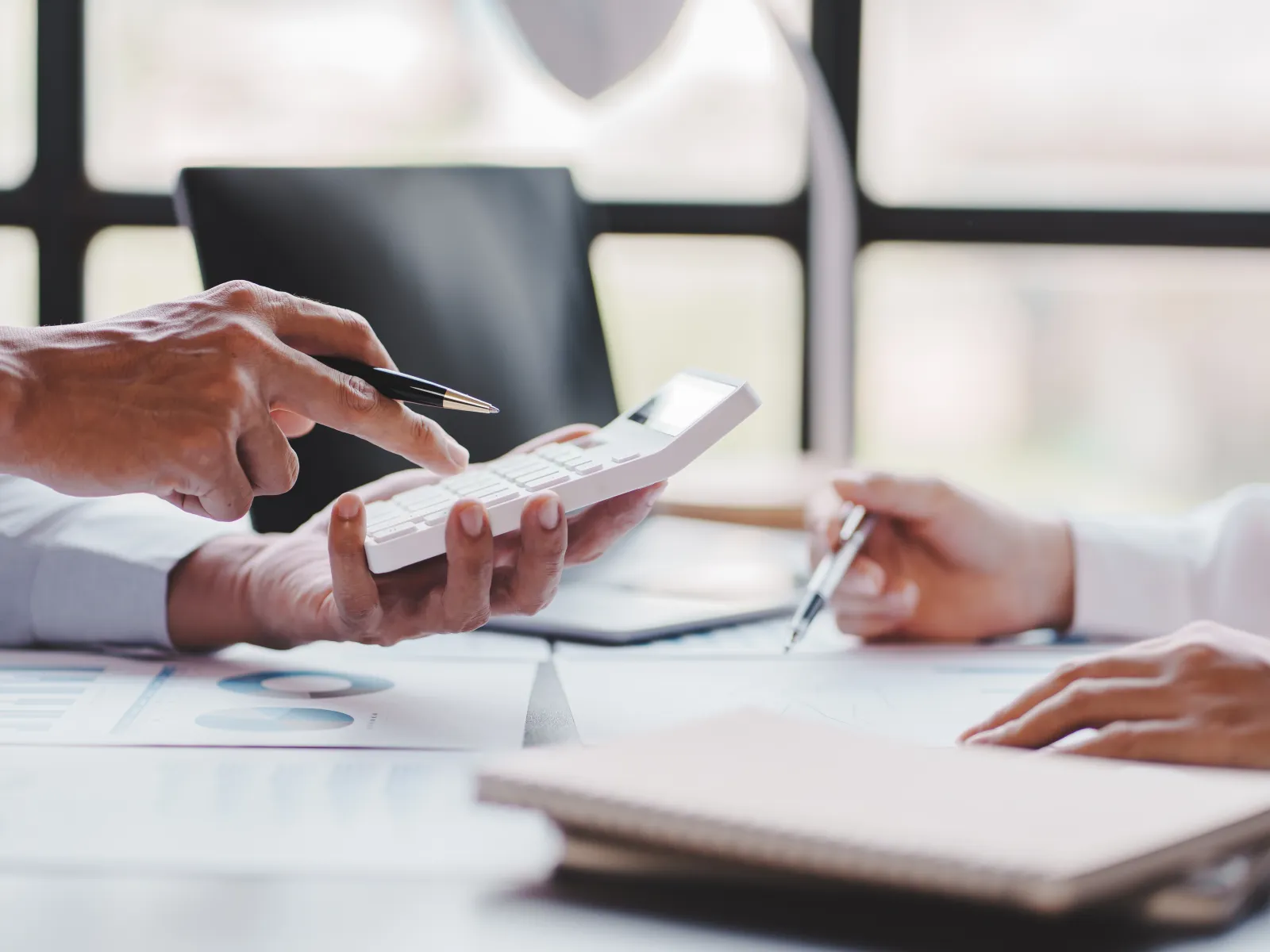 Two people using a calculator and writing notes while analyzing financial documents in a bright office.