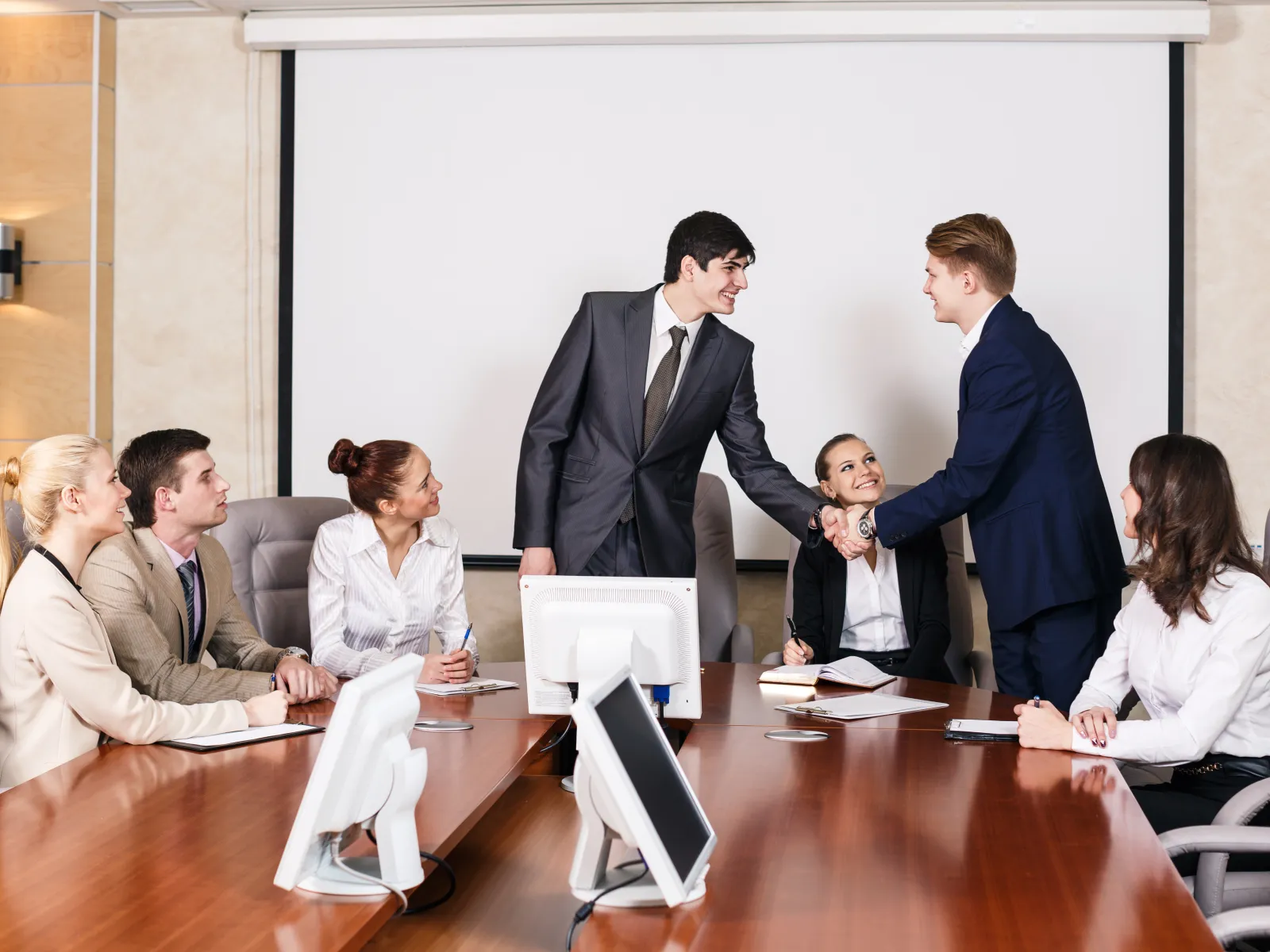 Business professionals in a meeting room shaking hands in front of a projector screen during discussion