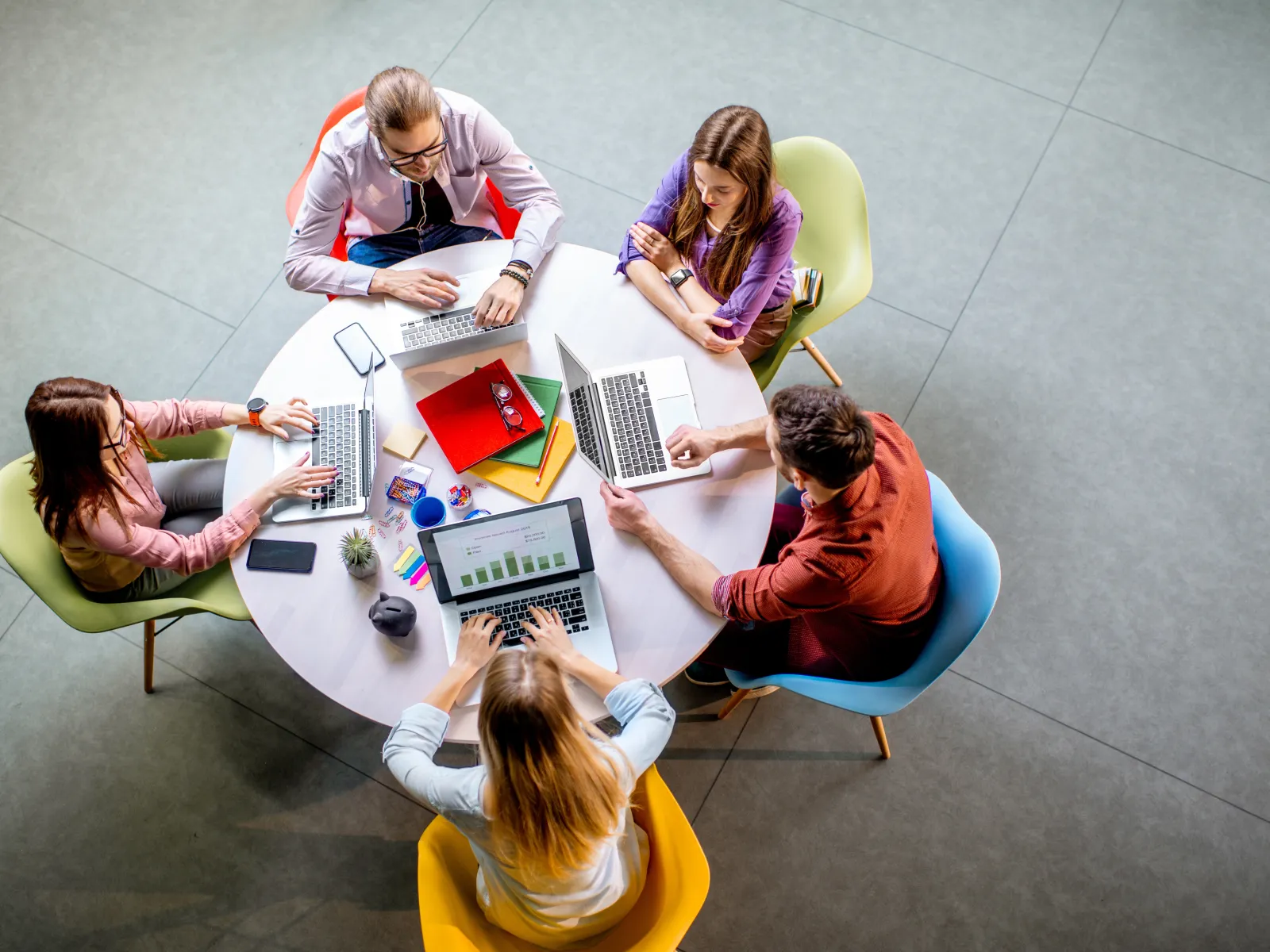 Top view of five people collaborating around a round table with laptops and colorful notebooks in a modern office.