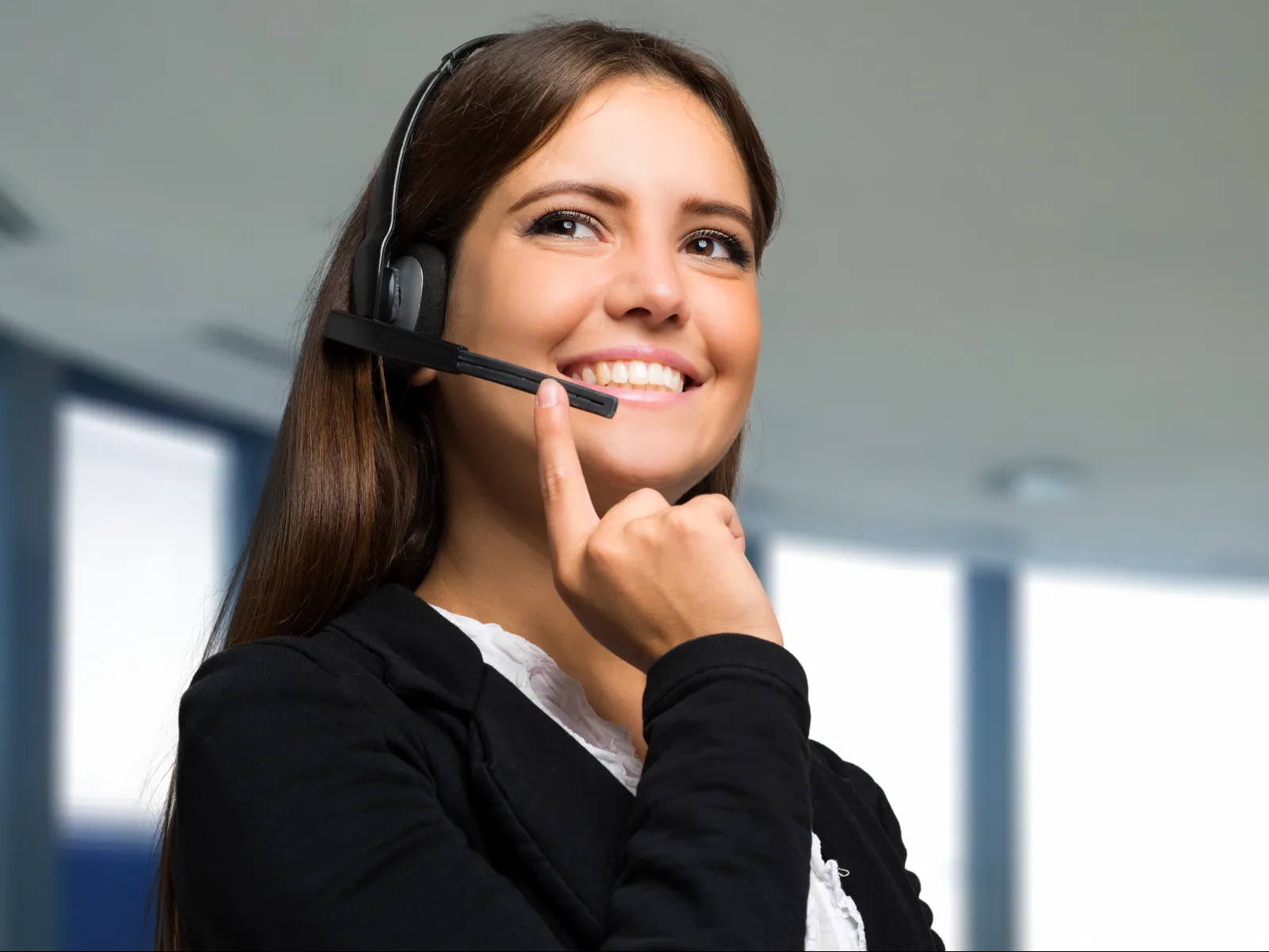 Smiling customer service woman wearing headset in modern office environment with bright windows.