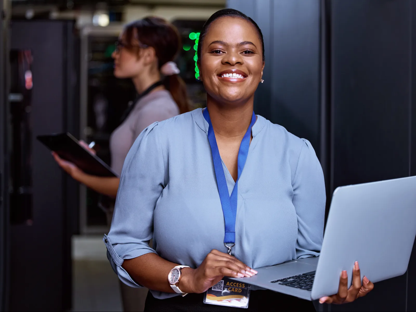Smiling woman holding a laptop in a data center, showcasing tech and teamwork.