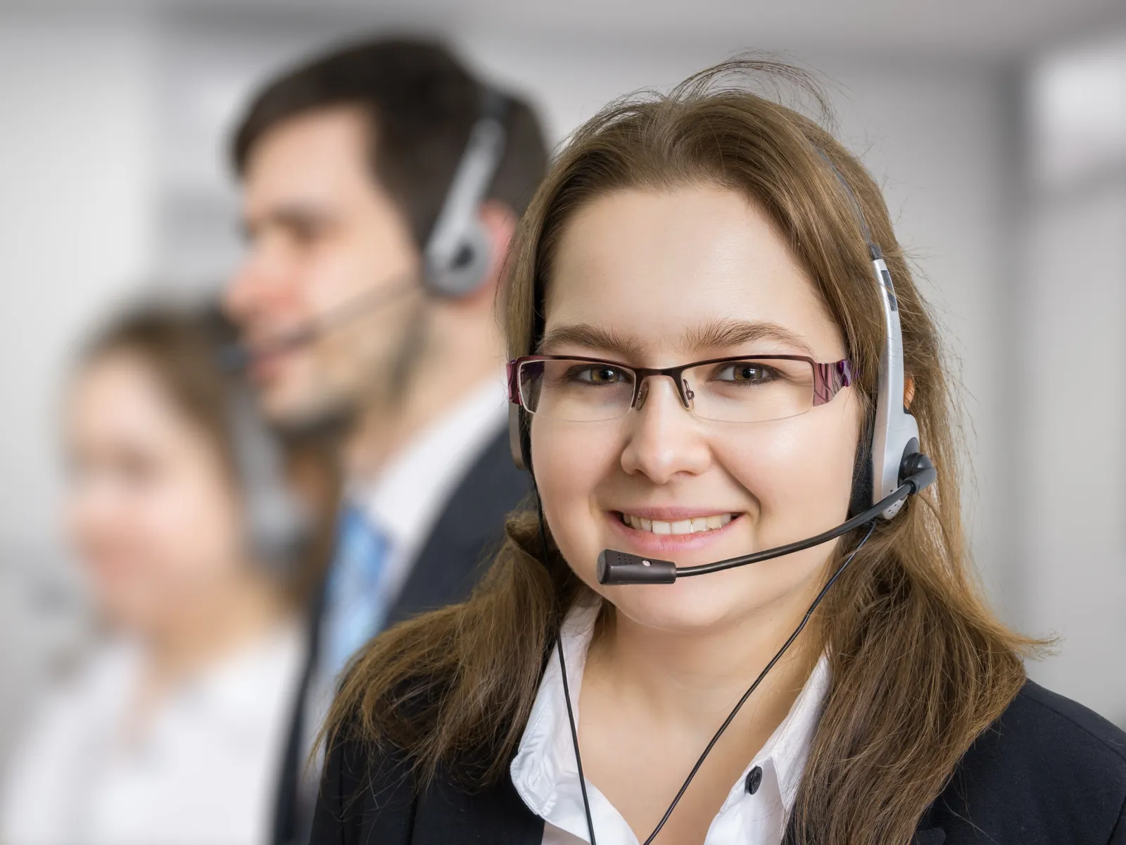 Smiling female customer service representative with headset in office, colleagues blurred in background