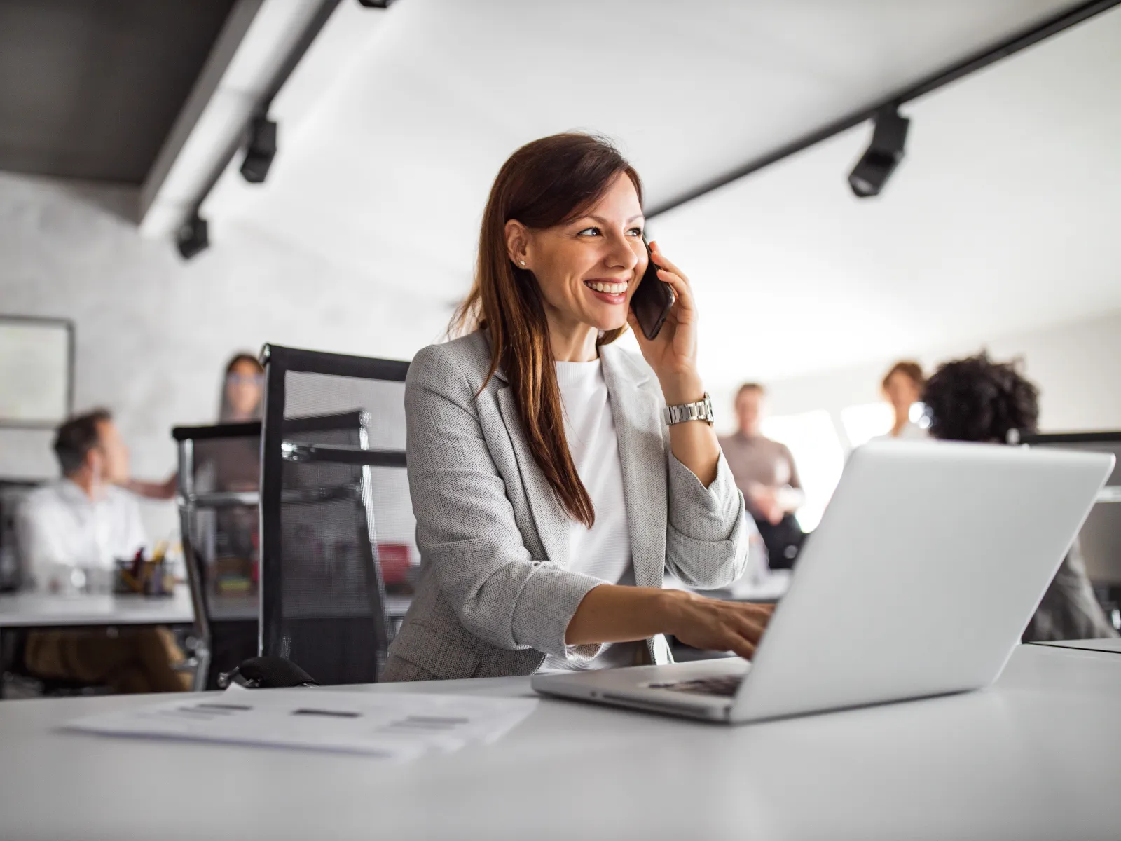 A businesswoman smiles while talking on the phone and working on her laptop in a modern office.
