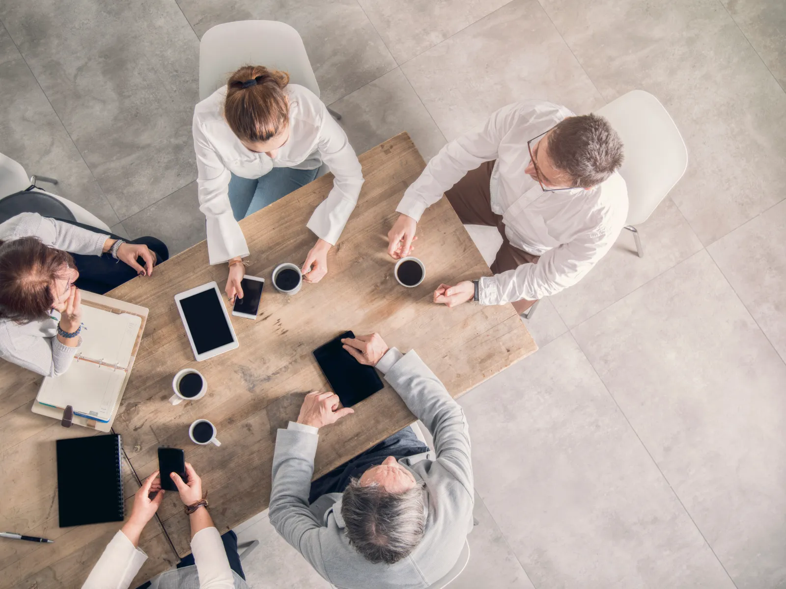 Overhead view of five professionals having a meeting around a wooden table with coffee and digital devices.
