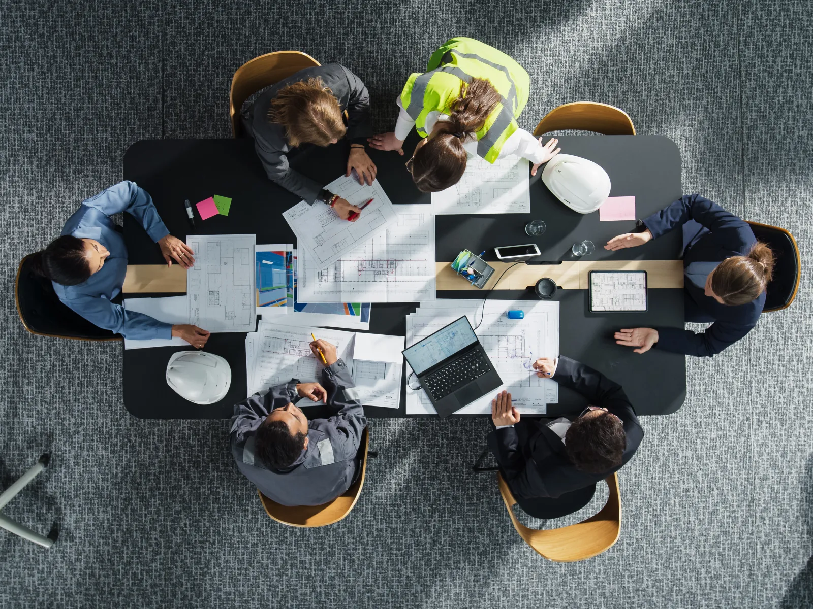 Overhead view of a diverse team collaborating on architectural blueprints and laptops at a modern office table.