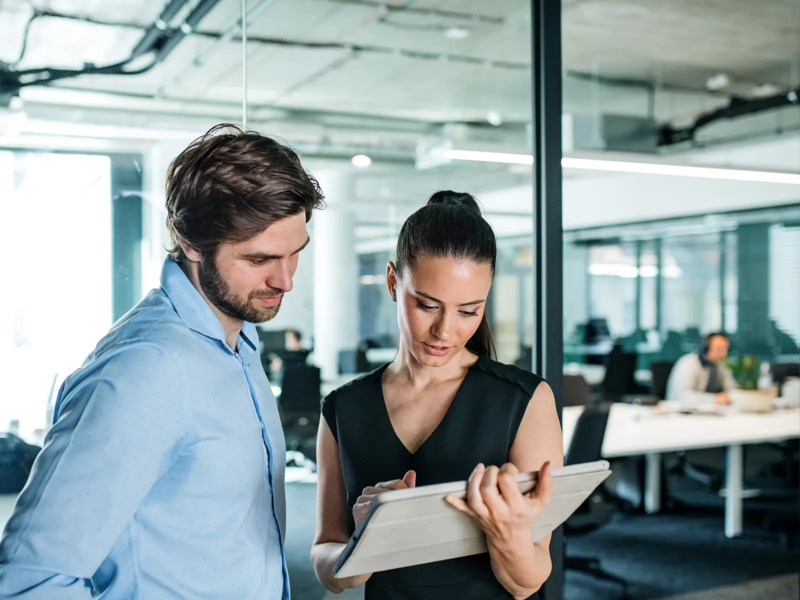 Two colleagues review information on a tablet together in a modern office workspace with glass walls.