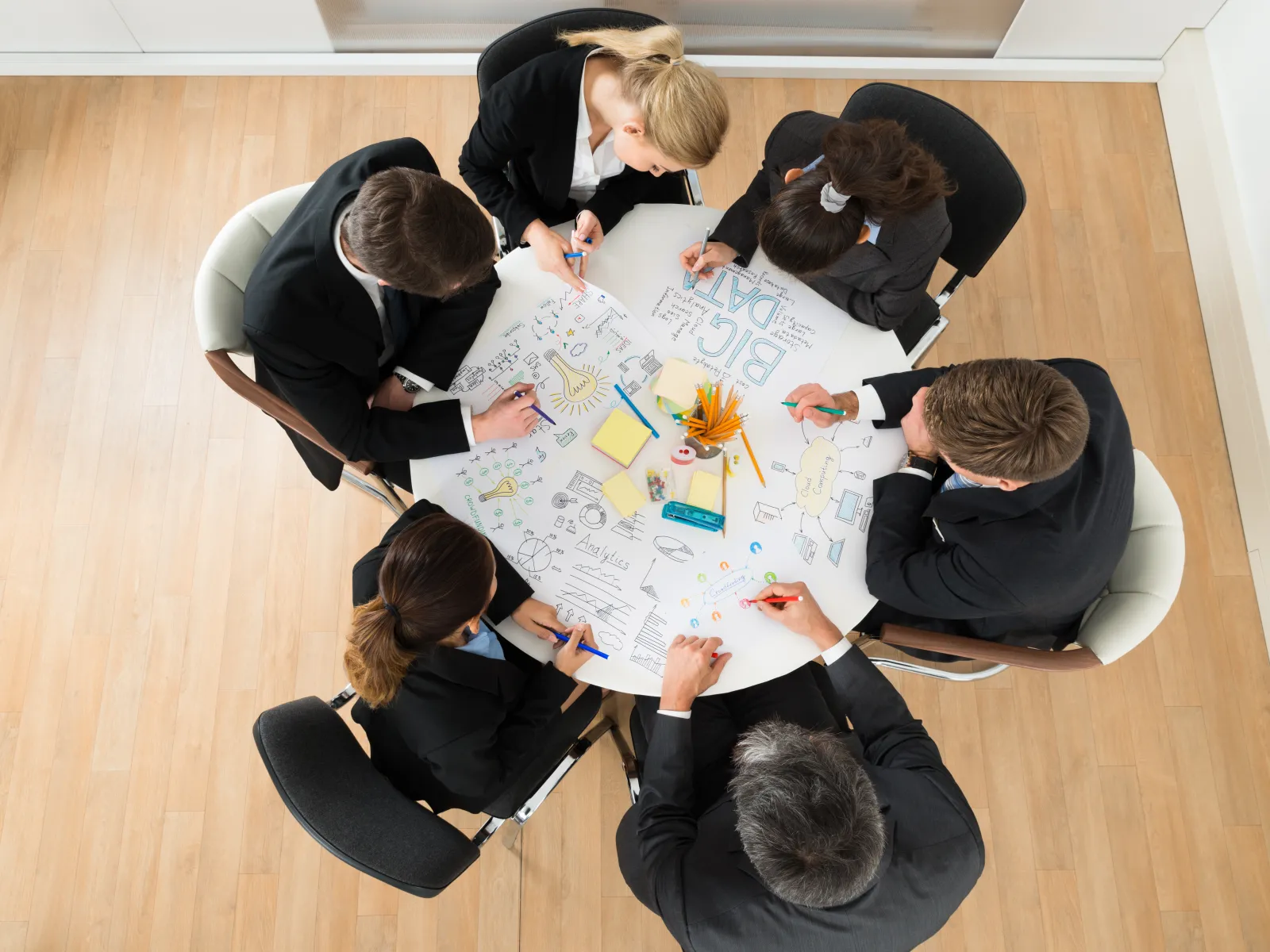 Overhead view of six professionals collaborating around a white table with colorful diagrams and notes.