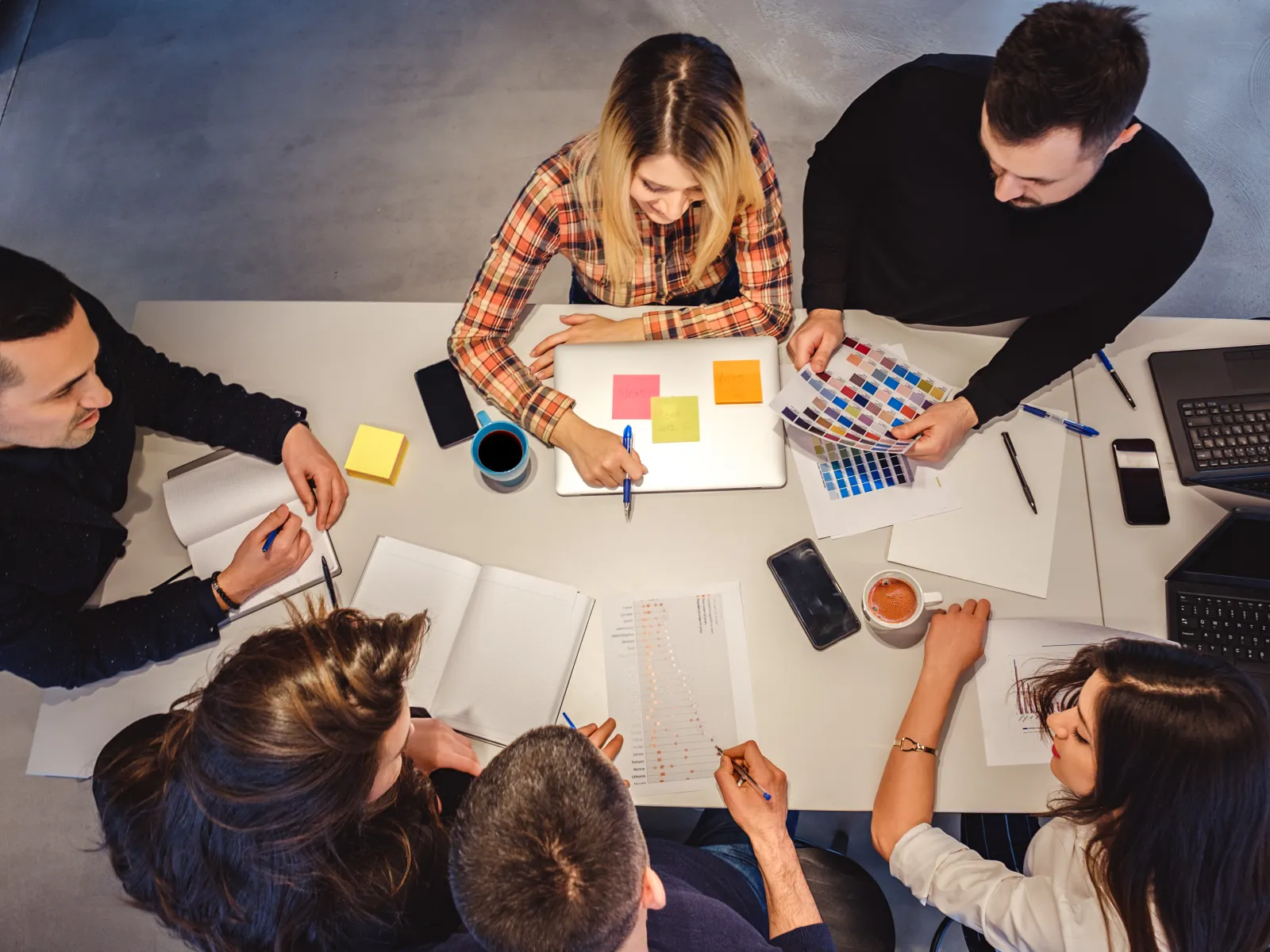 Top view of a team collaborating at a table with laptops, notes, color swatches, and coffee during a meeting.