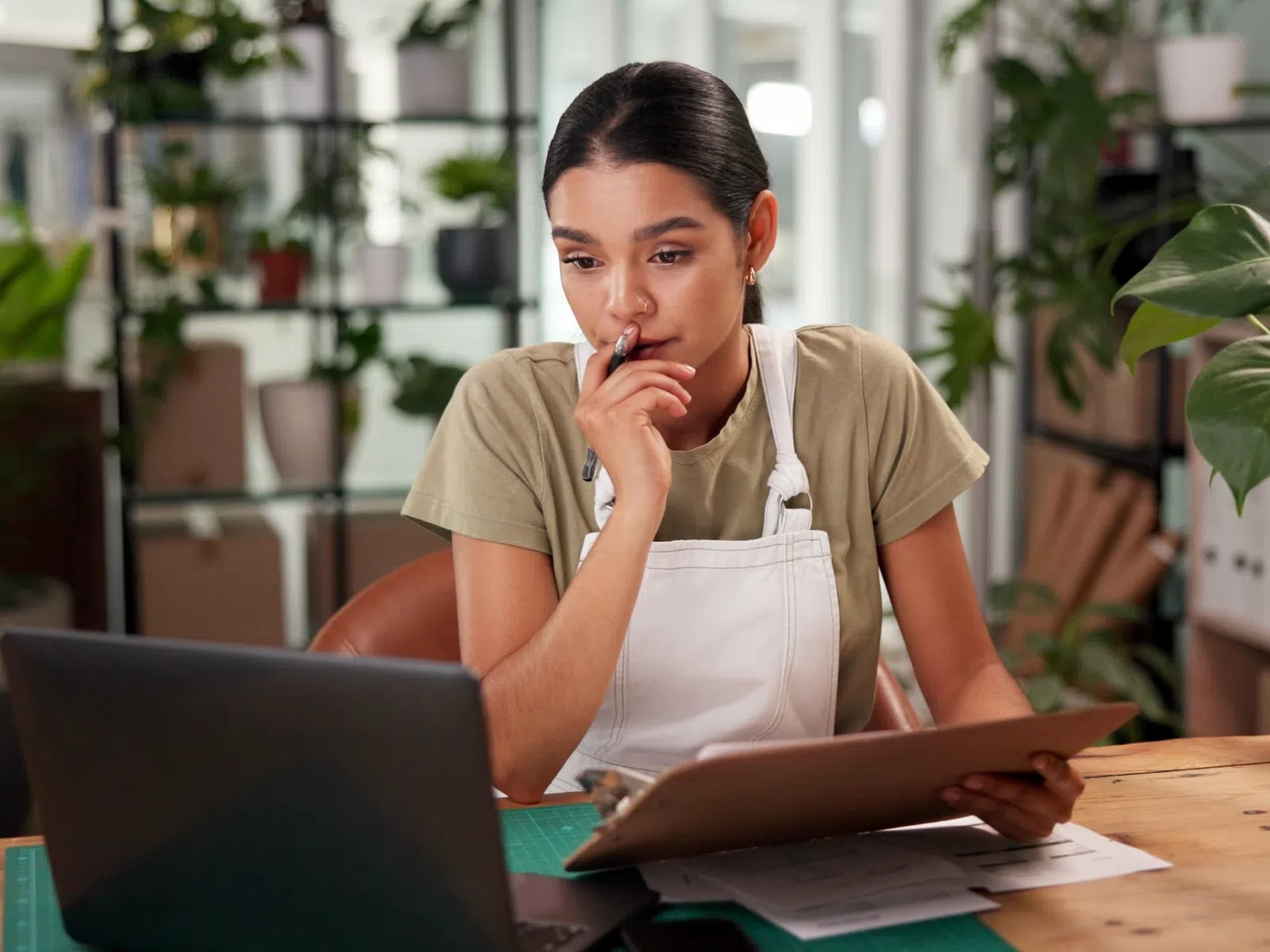 Young woman in apron working thoughtfully on laptop and tablet in a plant-filled creative workspace.