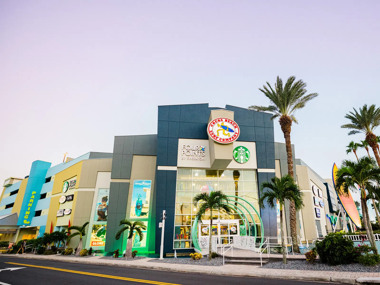 Exterior of a modern shopping center with palm trees, featuring Starbucks, Four Points and other stores at sunset.