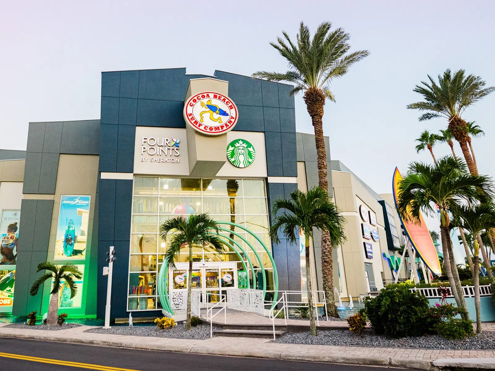 Exterior of the Cocoa Beach Surf Company in Cocoa Beach, Florida, a modern shopping center with palm trees, featuring Starbucks, Four Points and other stores at sunset.