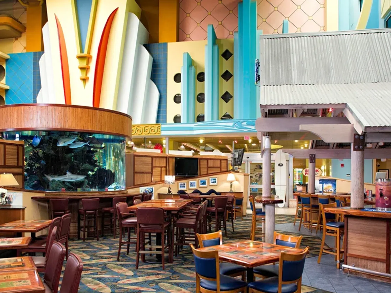 Colorful, retro-style restaurant interior with wooden tables, bar area, and large cylindrical fish tank centerpiece