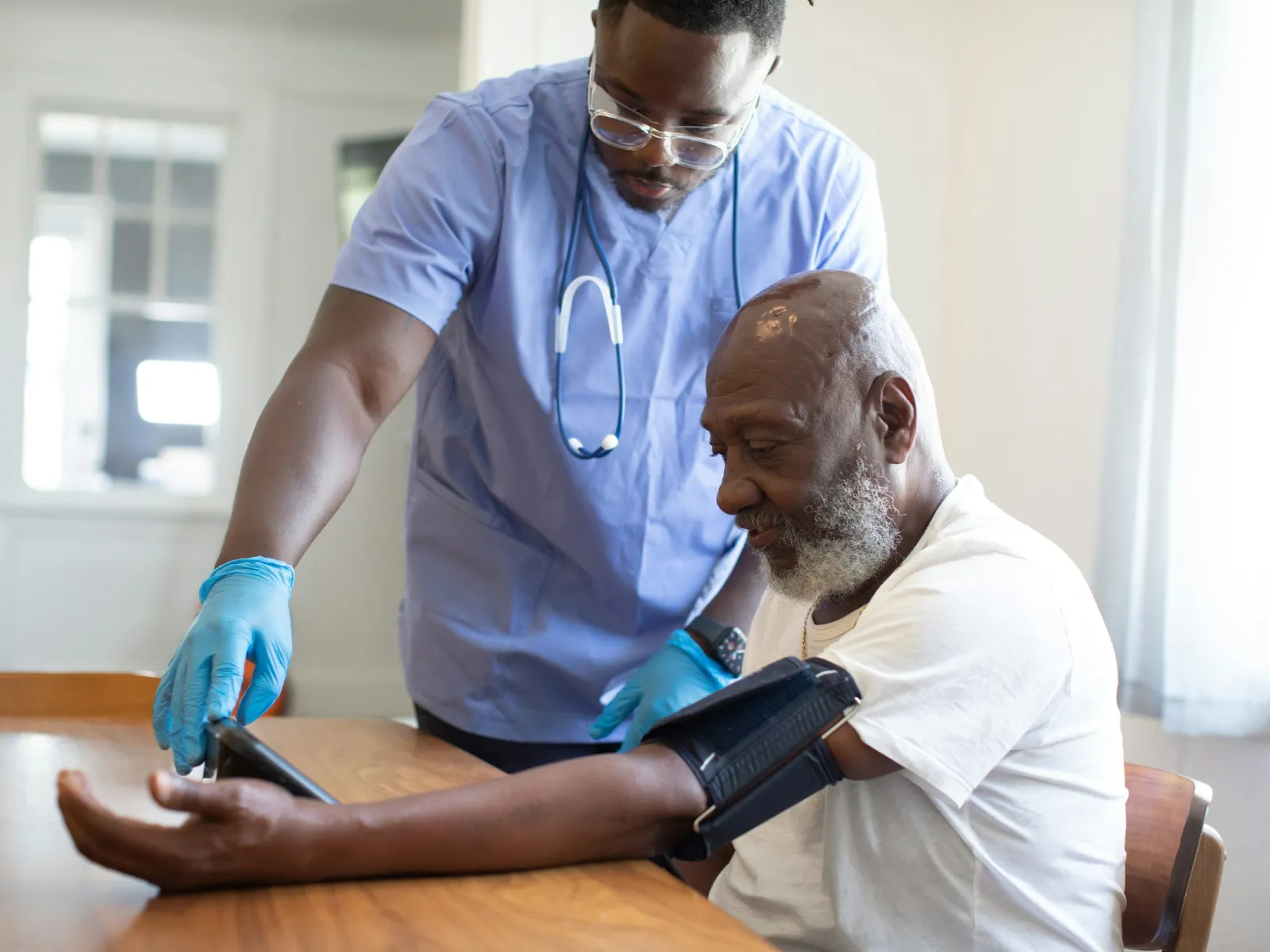 Healthcare worker measures blood pressure of elderly man at home with a digital cuff in a bright room.