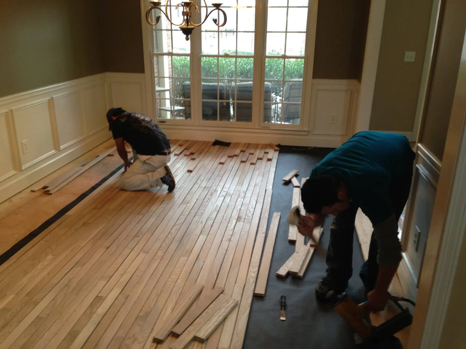 Two workers installing wooden flooring in a bright room with large windows and chandelier.