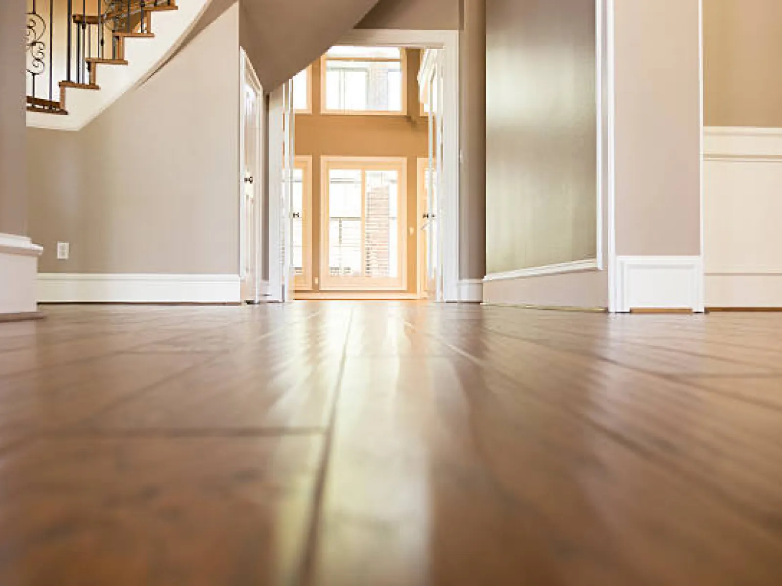 Bright empty hallway with polished hardwood floors, beige walls, white trim, and staircase with iron railing.