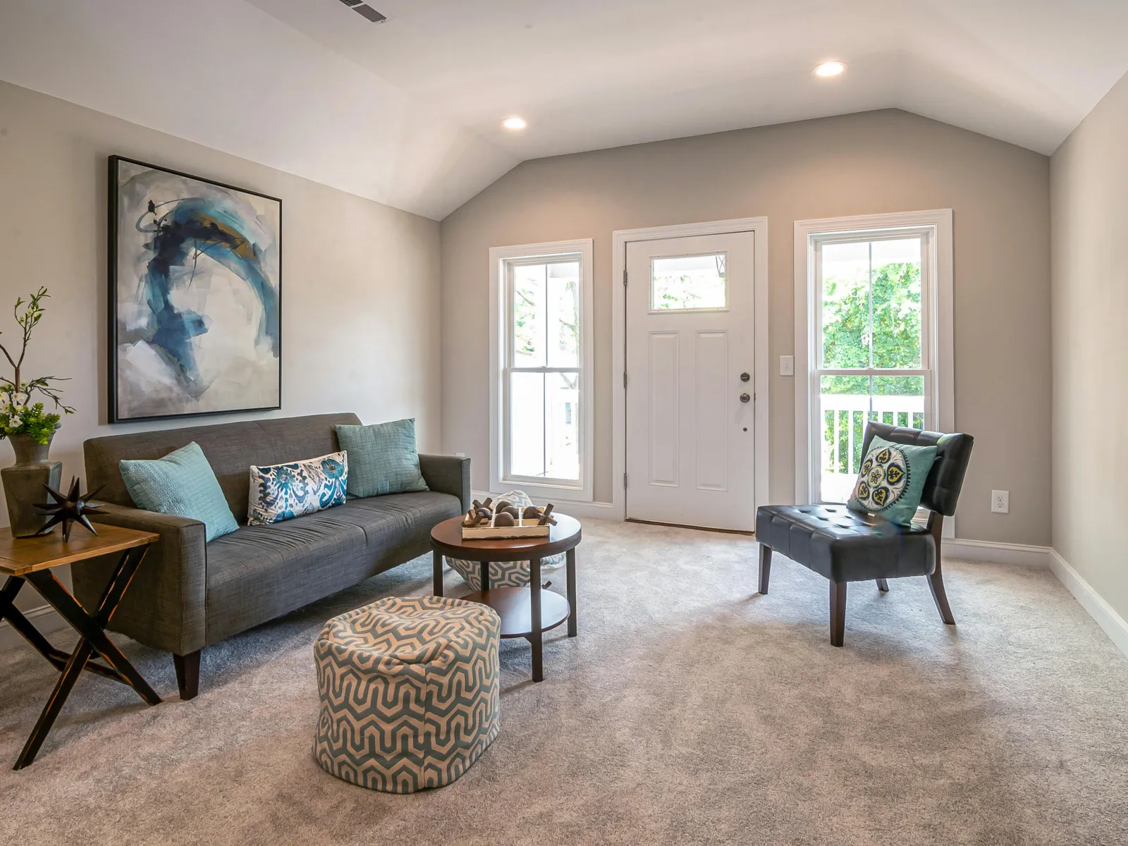 Bright living room with gray sofa, patterned ottoman, black armchair, abstract wall art, and beige walls.