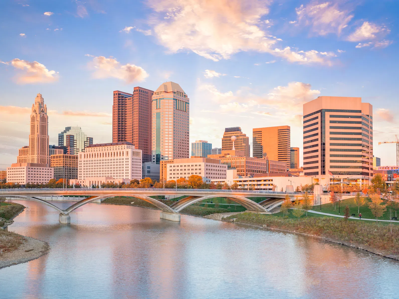 Columbus Ohio skyline at sunset with riverfront and arched bridge reflecting in the water under a colorful sky