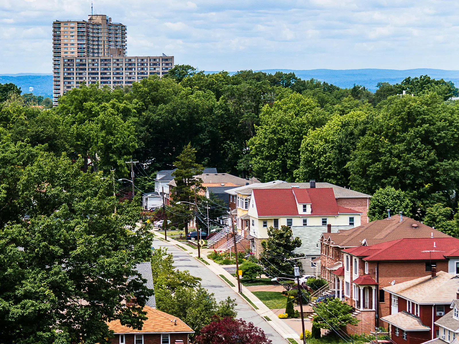 Residential neighborhood street with houses, trees, parked cars, and a tall apartment building in the background under a cloudy sky.