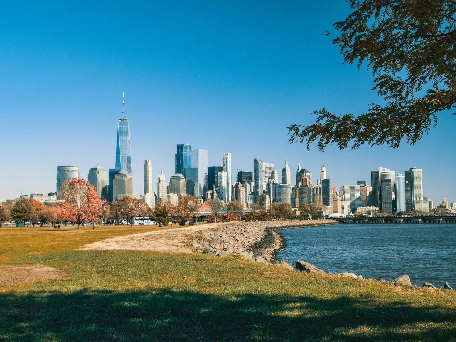 New York City skyline with One World Trade Center and autumn trees along waterfront park on a clear day