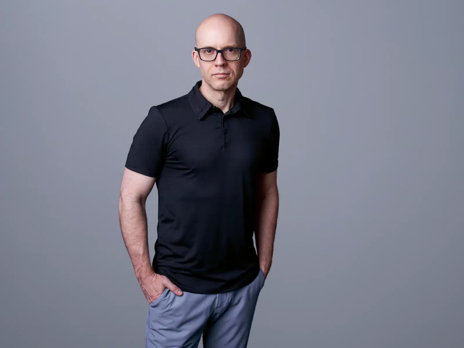 Bald man wearing glasses, black polo shirt, and gray pants standing with hands in pockets against gray background