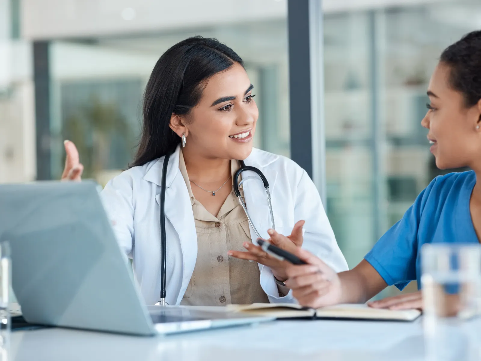 Female doctor and nurse discussing patient care over laptop and notes in hospital setting