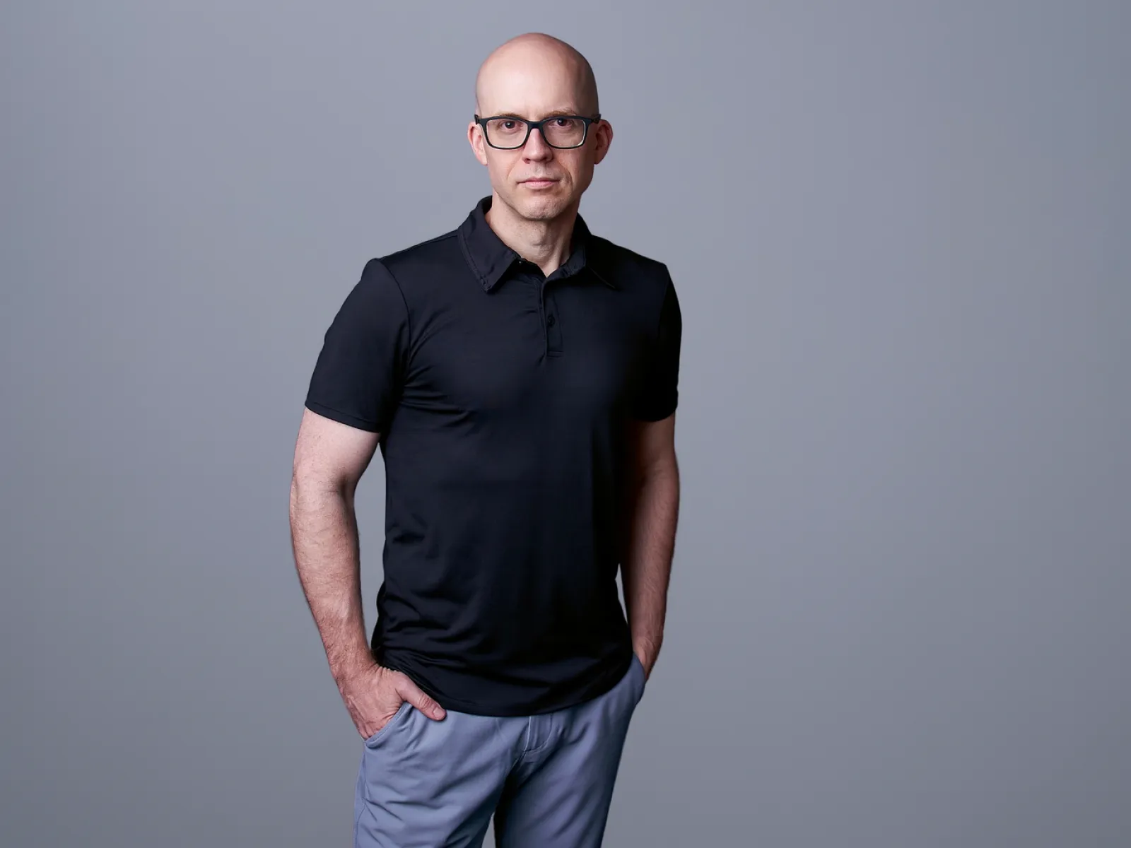 Bald man wearing glasses black polo shirt and gray pants standing with hands in pockets on gray background