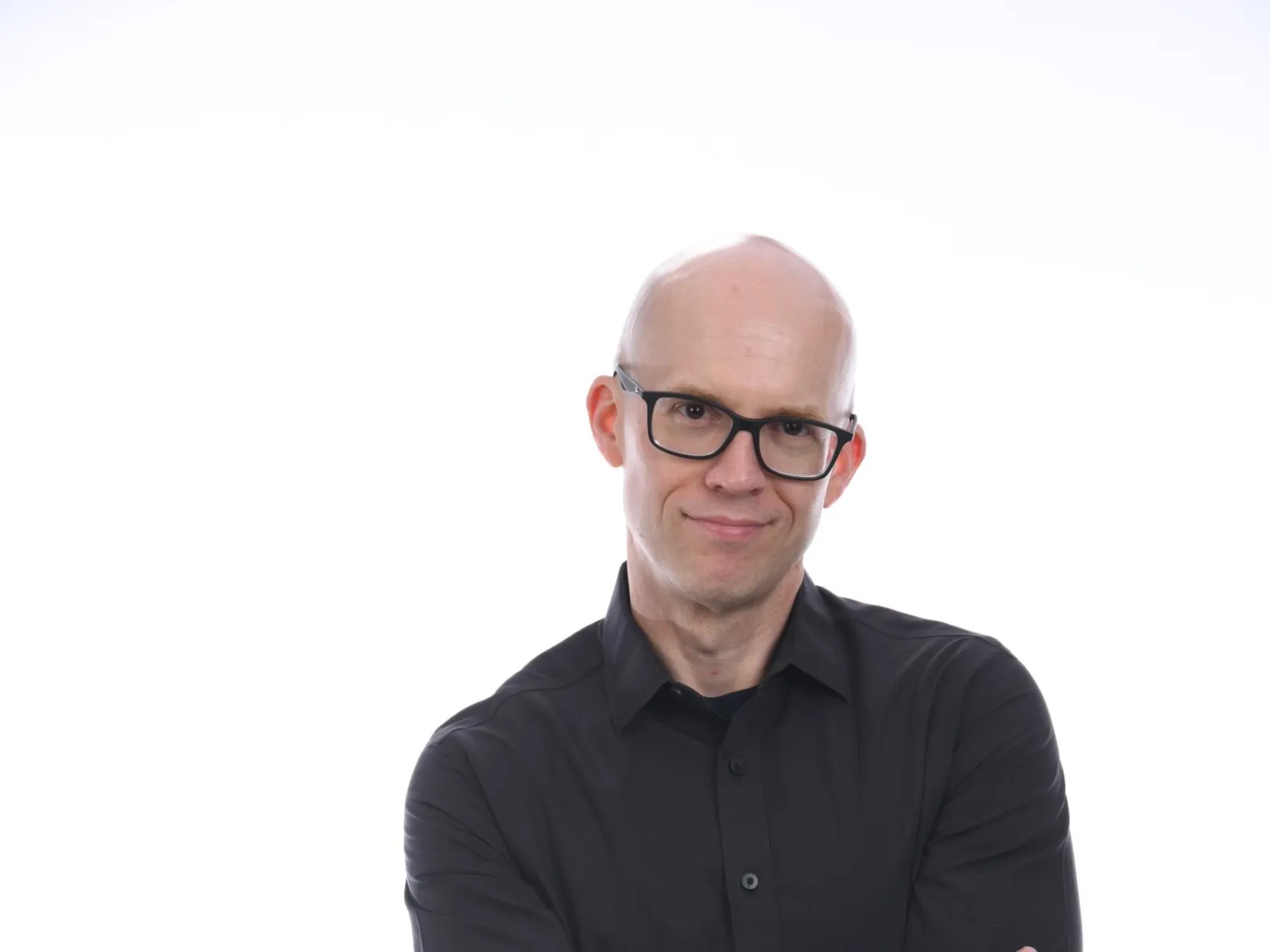 Bald man wearing glasses and black shirt with arms crossed against white background.