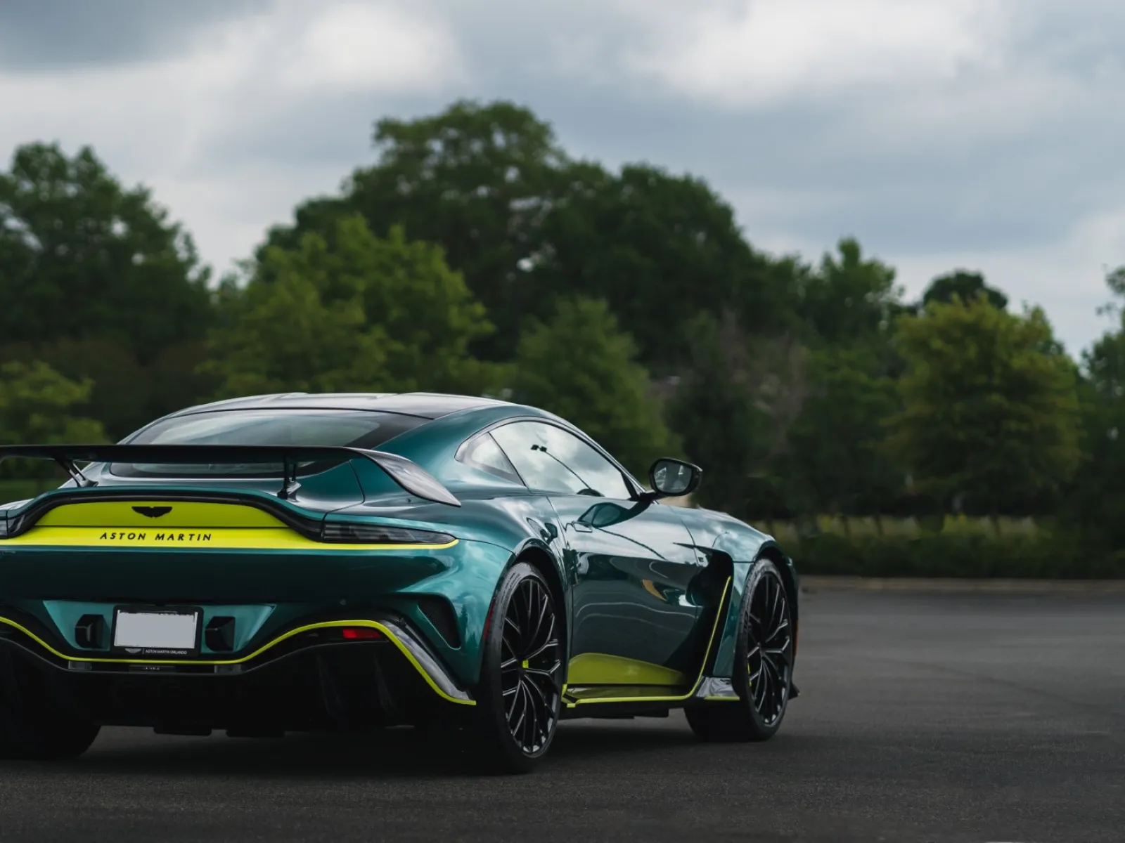 Green Aston Martin sports car with yellow accents parked on asphalt with trees in background under a cloudy sky