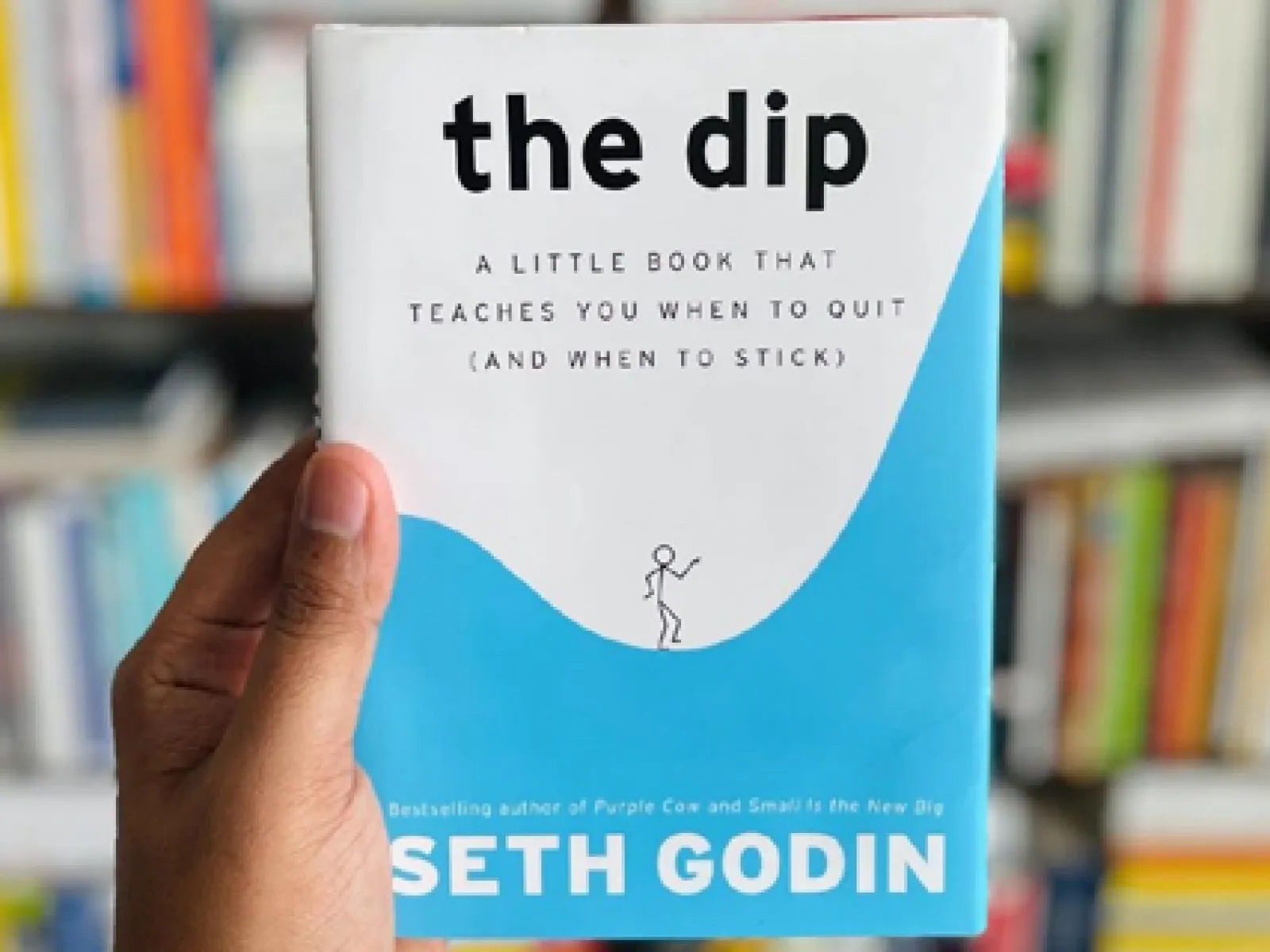 Hand holding the book 'The Dip' by Seth Godin with bookshelf blurred in the background.