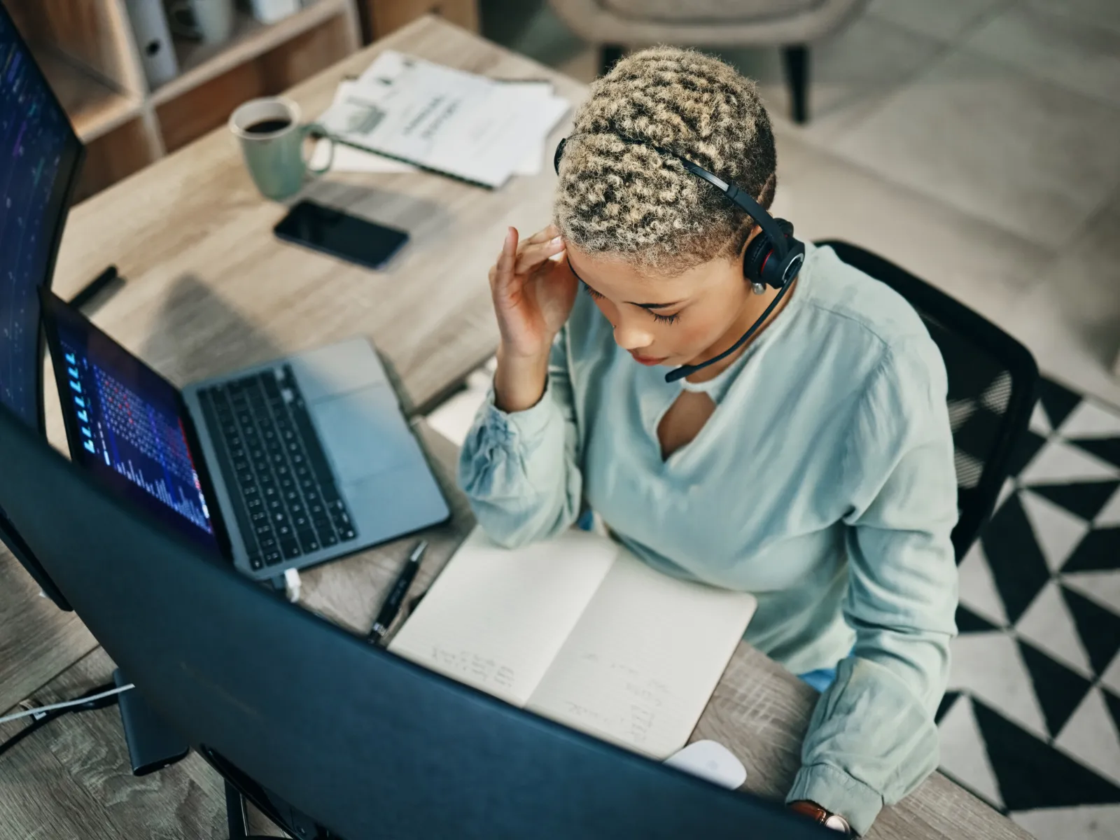 Overhead view of a woman wearing a headset working at a desk with dual monitors and a notebook in a home office.