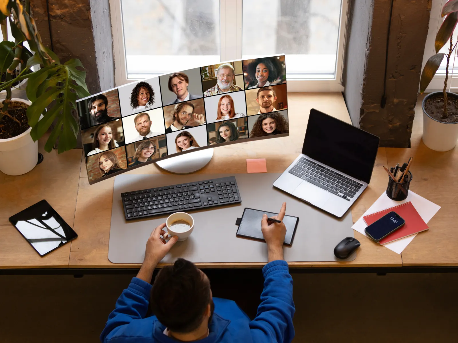 Person at desk using digital pen and laptop for a remote video meeting with multiple participants on curved monitor.