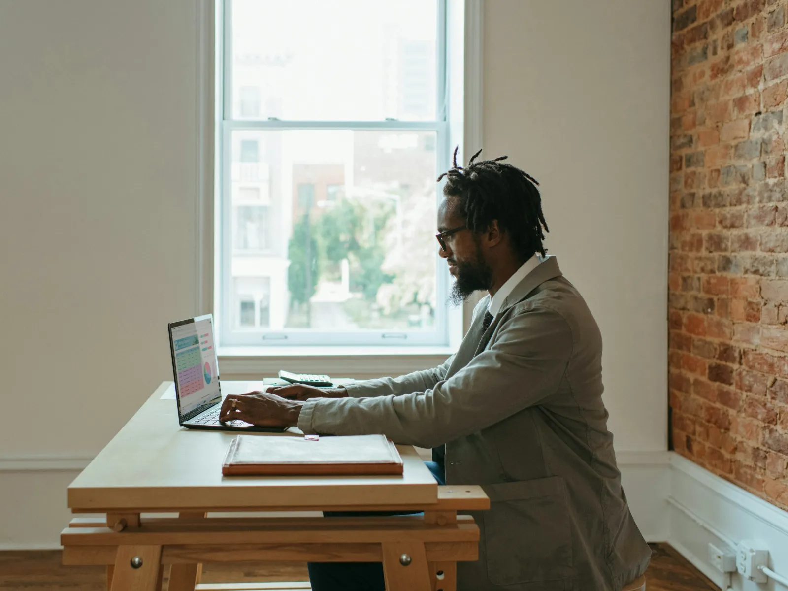 Man in glasses and blazer working on laptop at wooden desk near window in bright modern office.