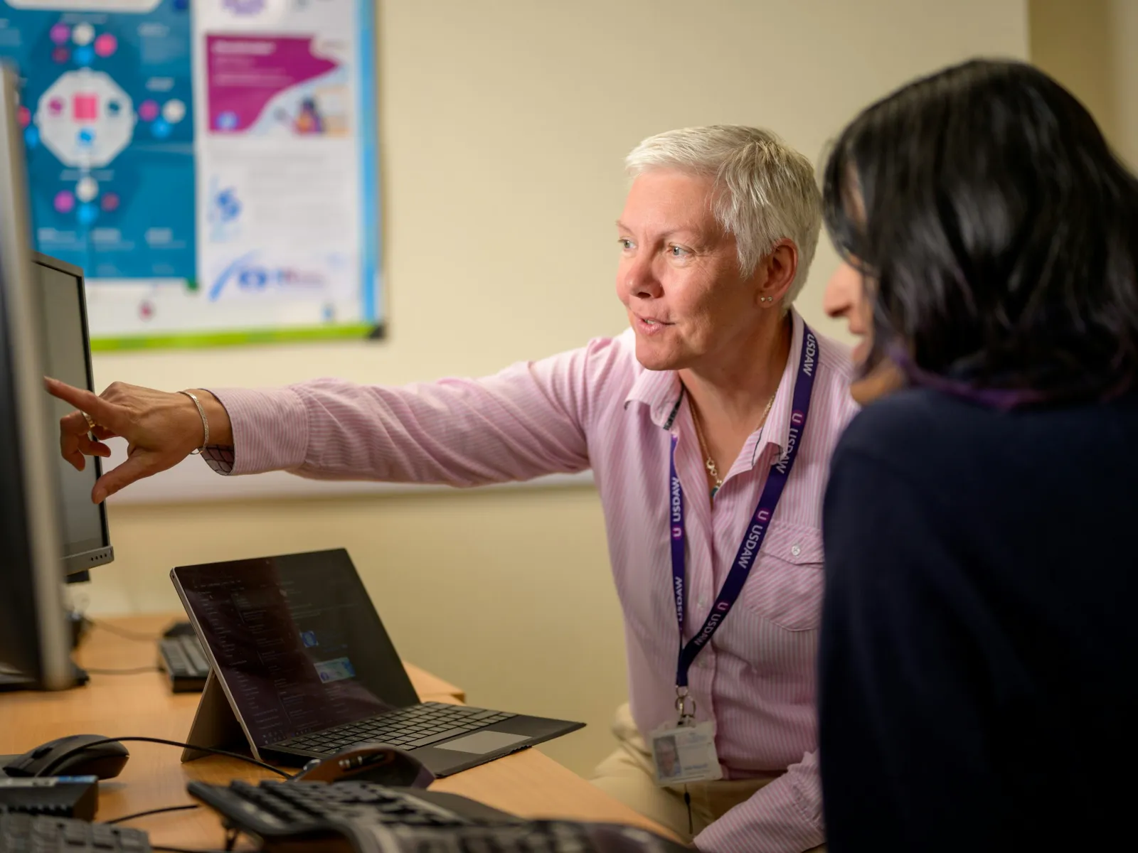 Senior woman instructing a colleague on computer use in an office setting with laptops and monitors.