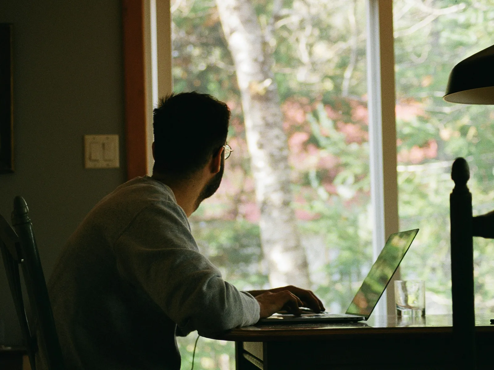 Man working on a laptop at a wooden table with a glass of water near a large window showing a green leafy background.
