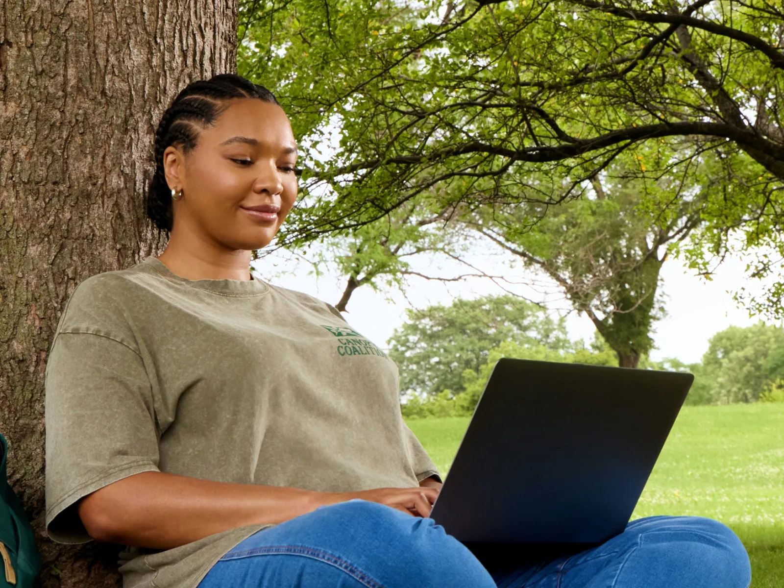Woman sitting against a tree working on a laptop outdoors in a green park with books and coffee nearby