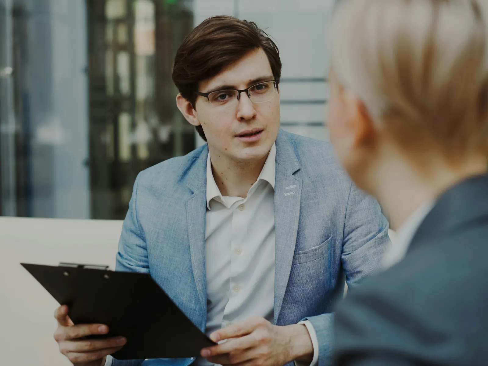 Young man in glasses and blue blazer holding clipboard talking to a woman in a professional office setting