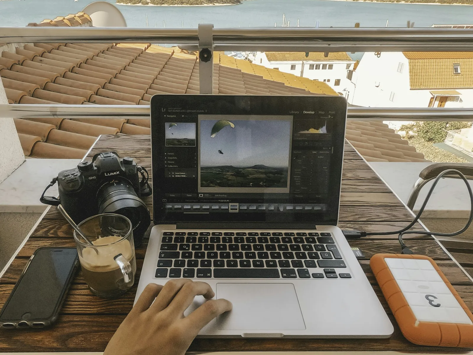 Person editing photos on a laptop at a wooden table with camera, coffee, external drive, overlooking sea and hills.