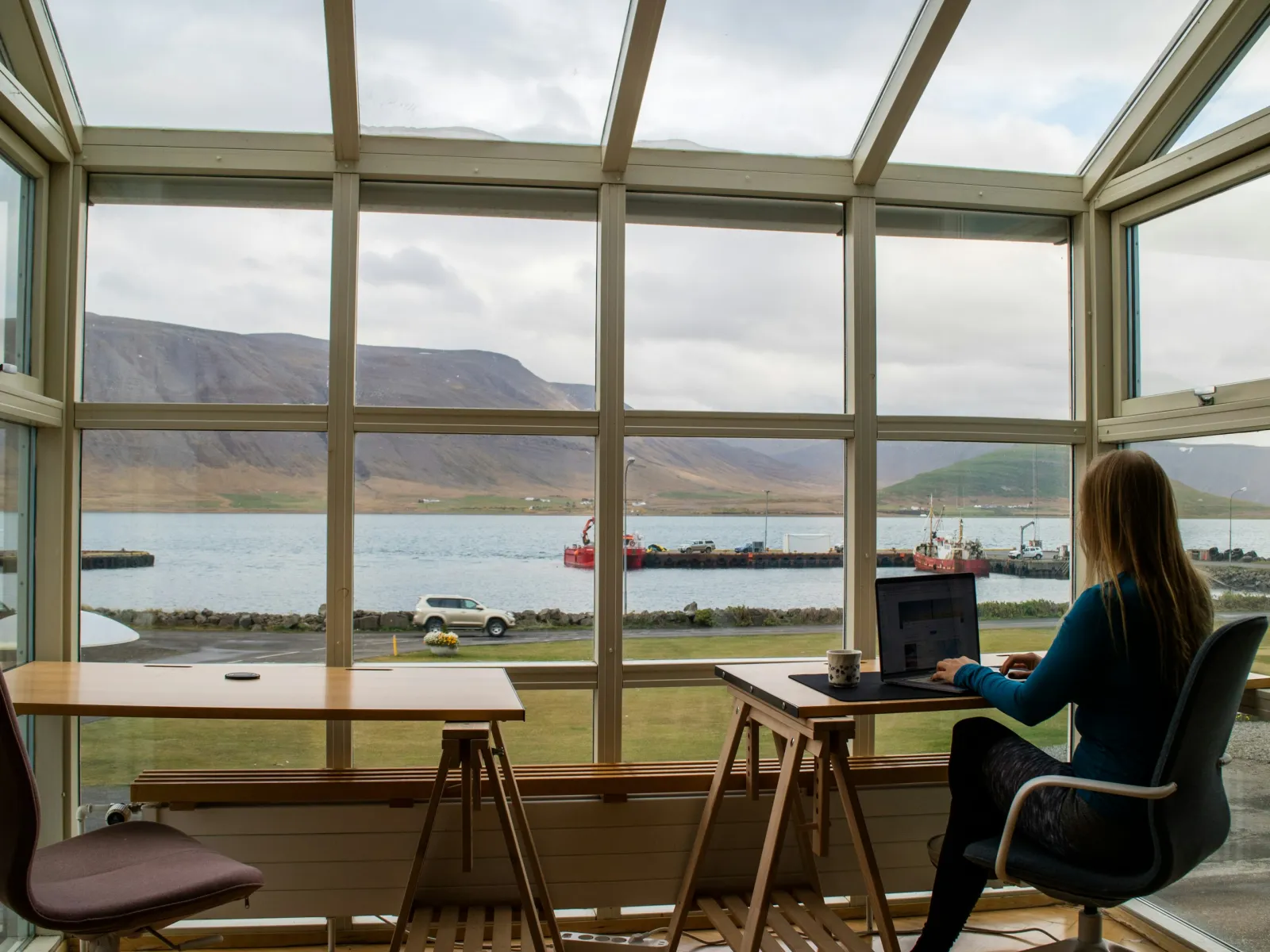 Woman working on laptop in a glass room with a scenic bay and mountains view outside under cloudy sky