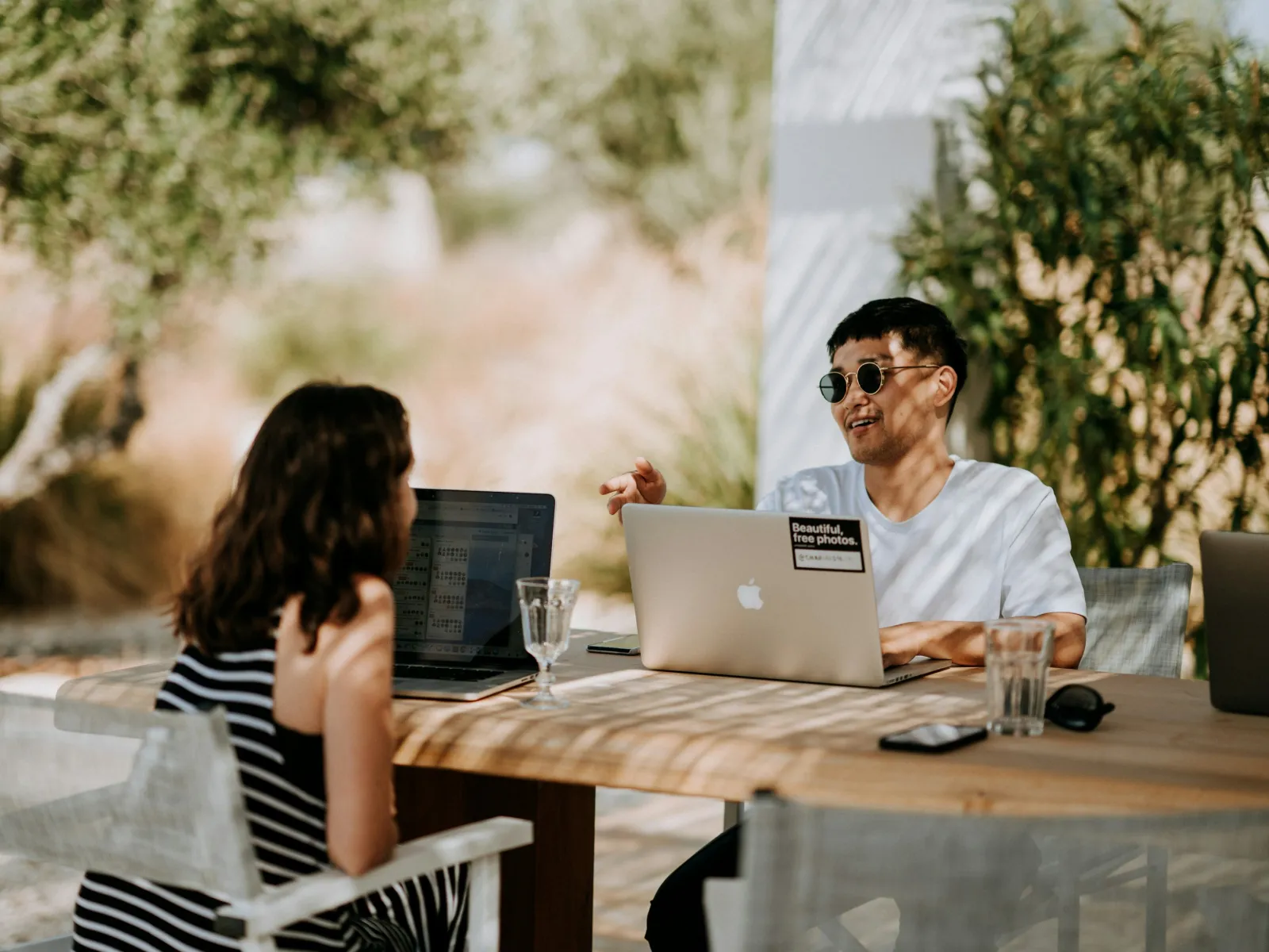 Two people having a casual outdoor meeting with laptops at a wooden table surrounded by greenery.