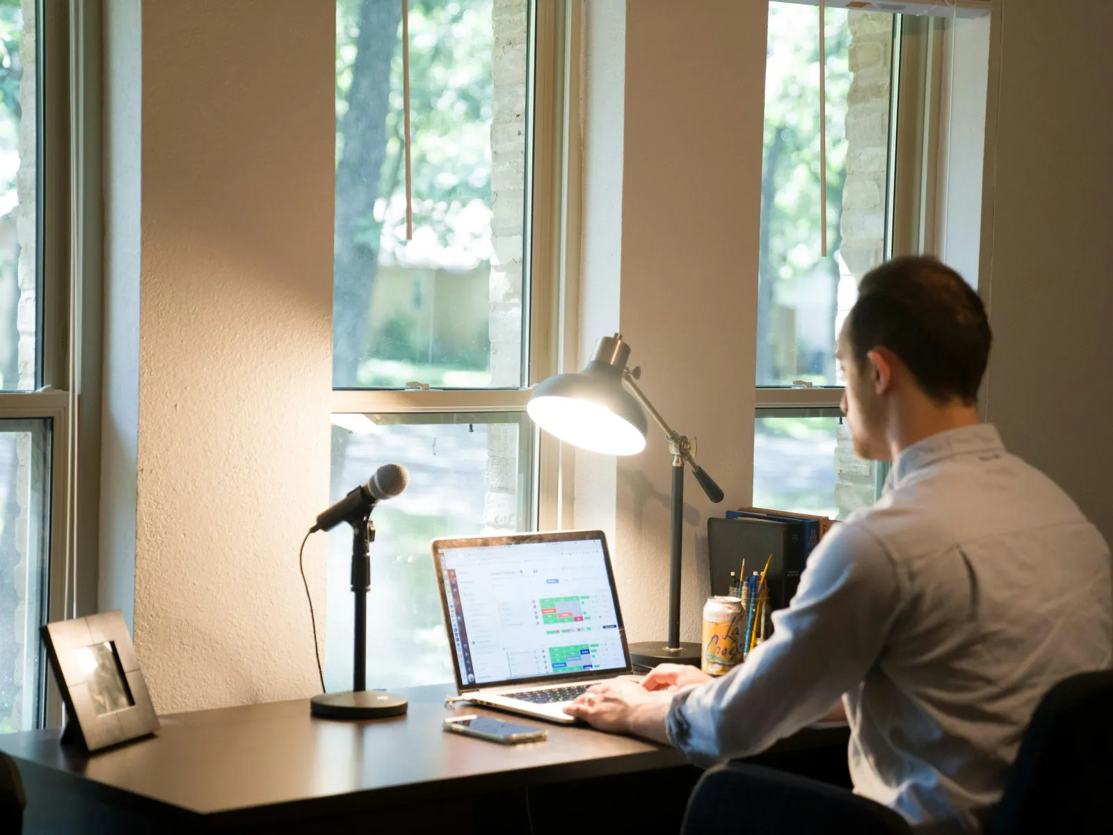 Man working on laptop at a desk with microphone, lamp, drink, and large windows letting in natural light.