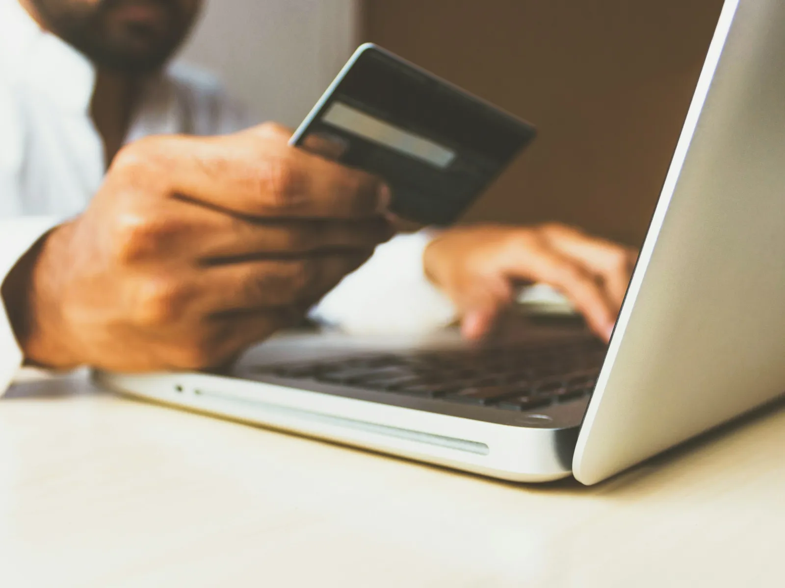 Man holding credit card and using laptop for online payment on a light wooden desk