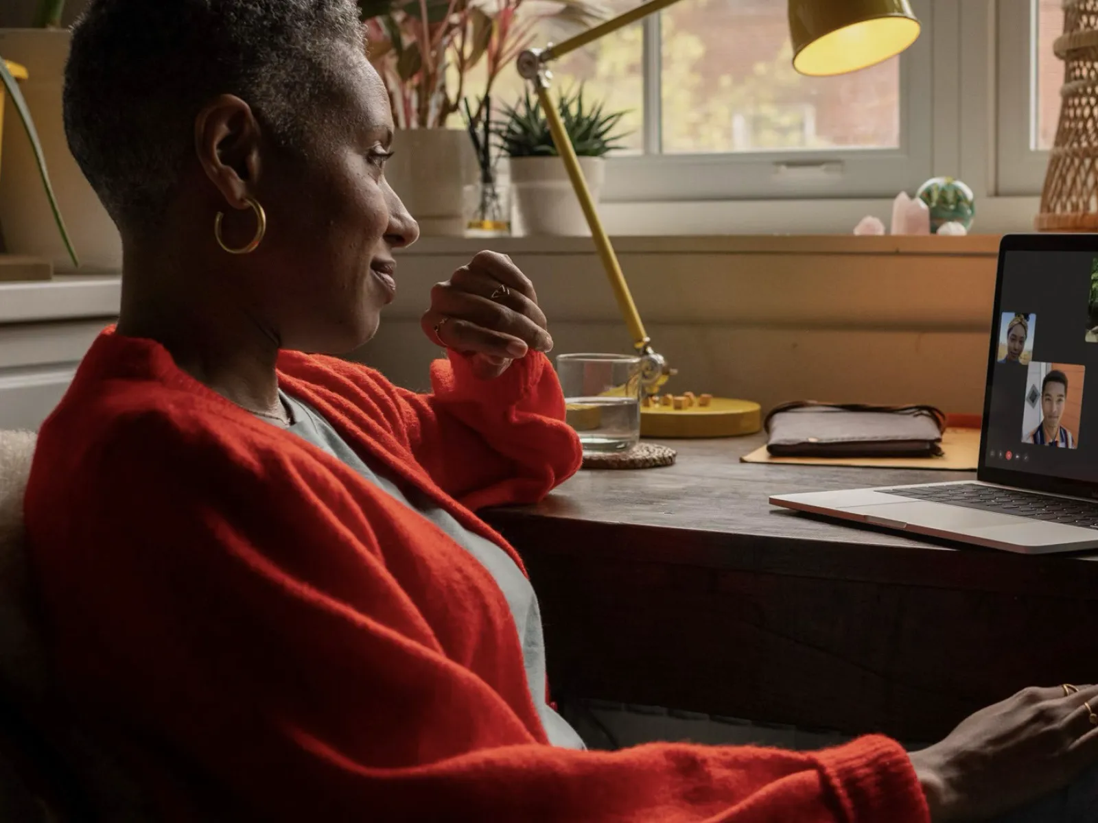 Woman wearing a red sweater video chatting with four people on a laptop in a cozy home office setting.