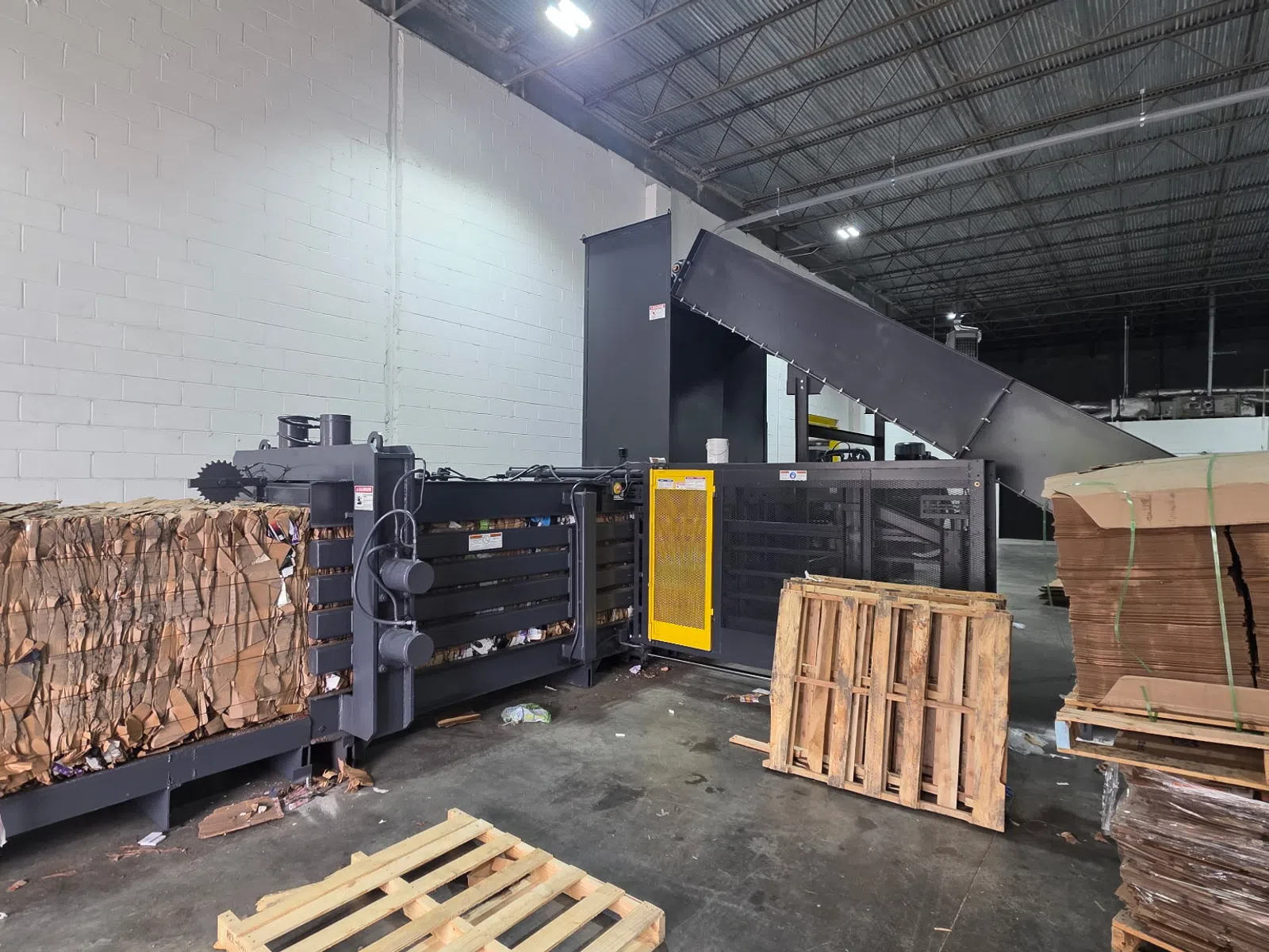 Industrial cardboard baler compressing cardboard waste into dense bales inside recycling warehouse with pallets.