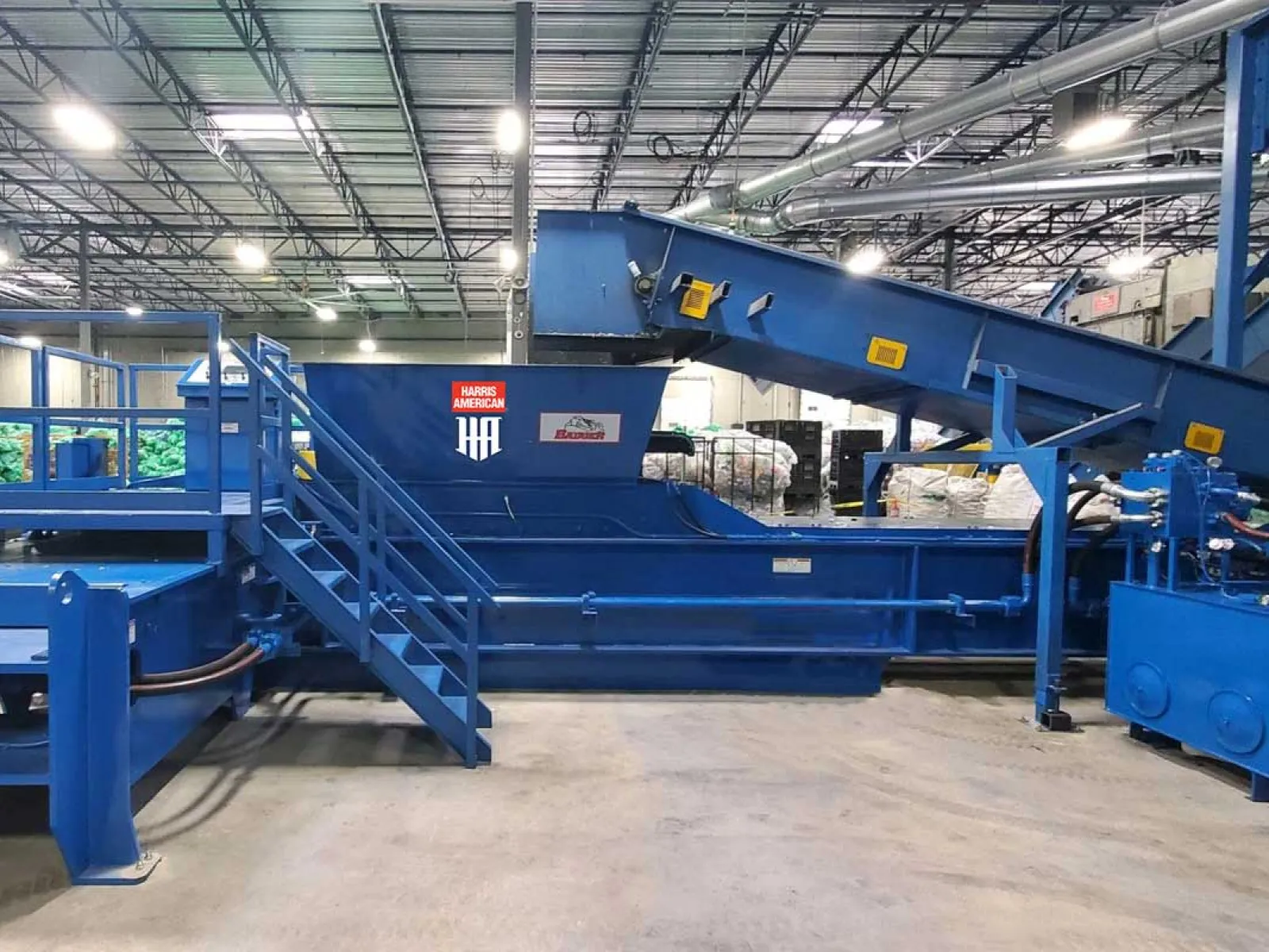 Large blue industrial conveyor system inside a warehouse with stairs and machinery under bright lighting