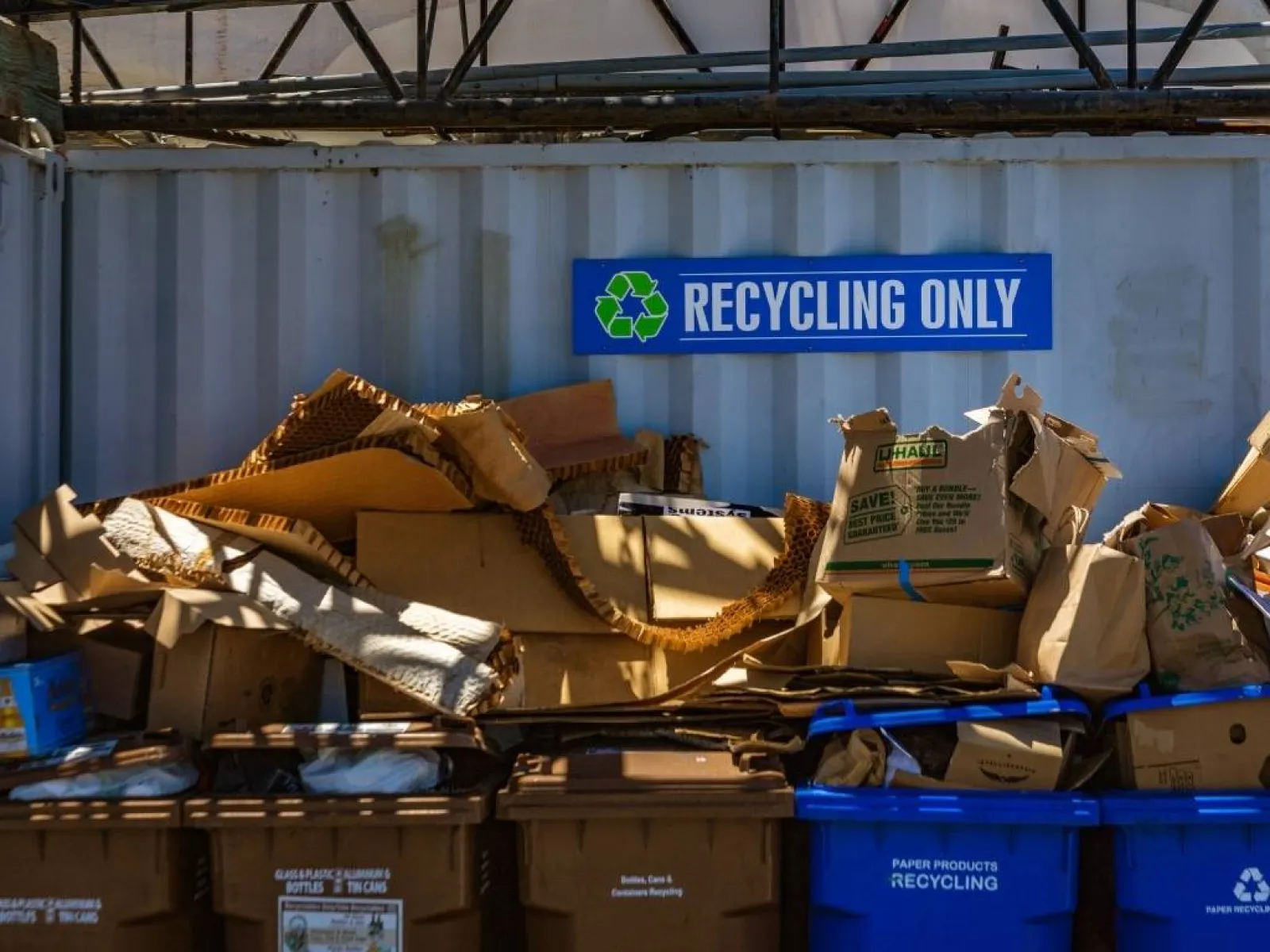Recycling station with blue bins and cardboard boxes under a container wall with a recycling only sign.