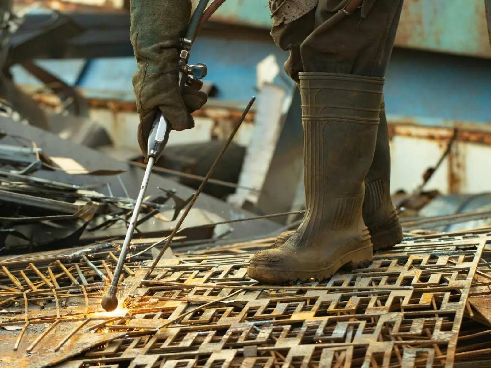 Worker wearing protective gloves and boots using a cutting torch on metal grates in construction site.