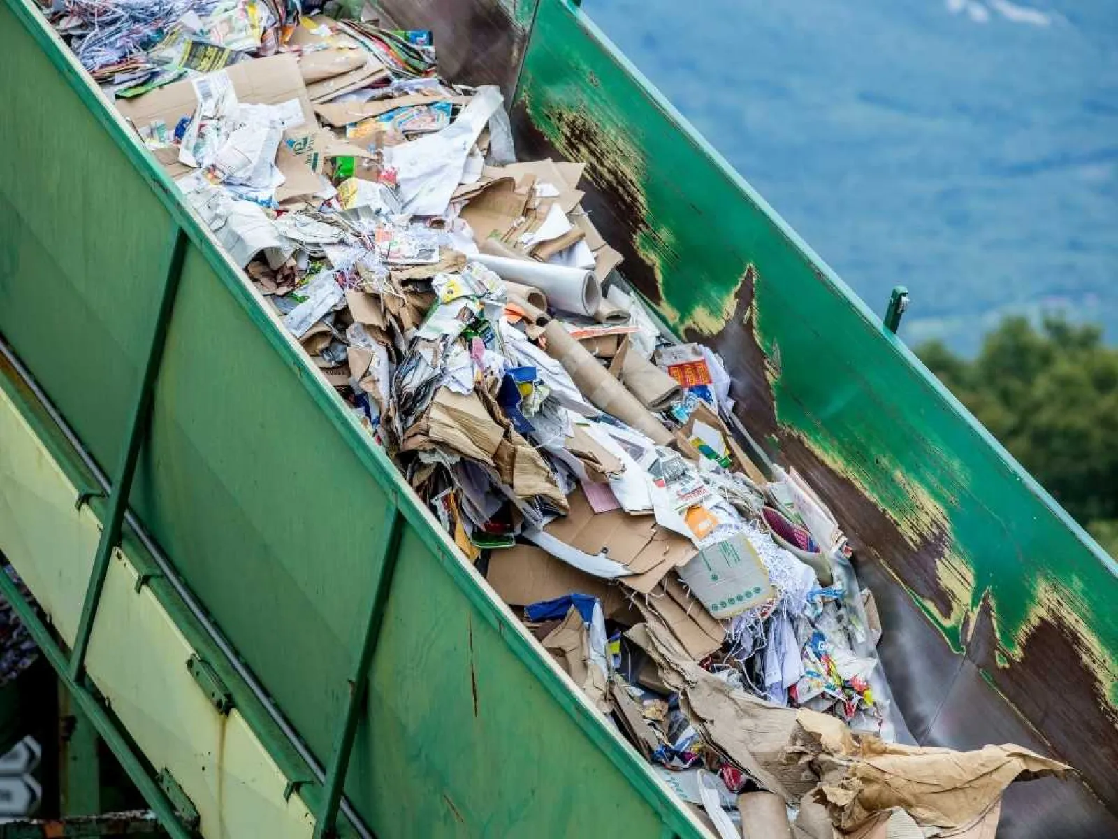 Large green dumpster filled with mixed recyclable cardboard, paper, and packaging waste outdoors.